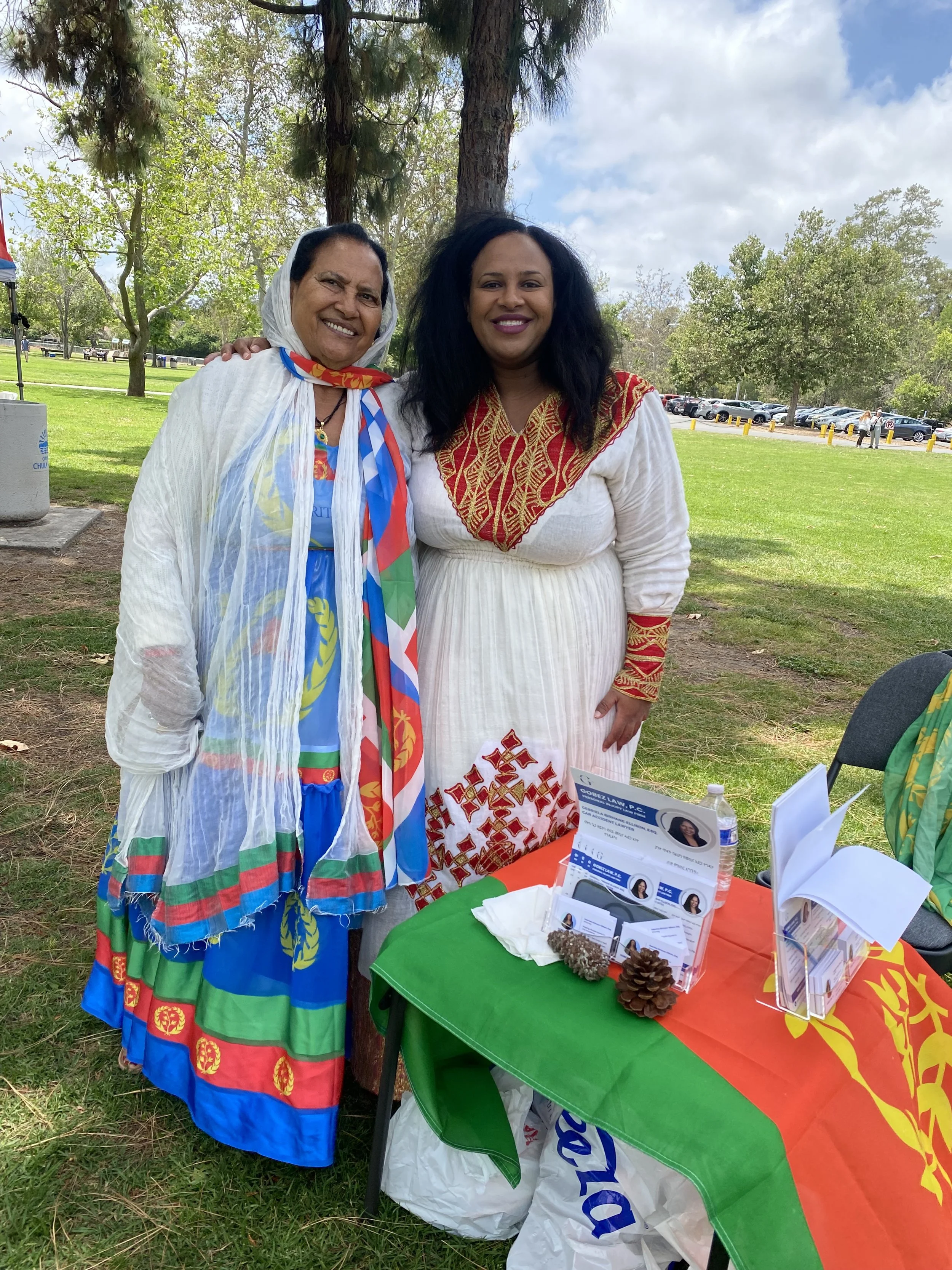 Two women in traditional Ethiopian attire standing outdoors at a cultural event, smiling, with a table displaying pamphlets and a flag draped over it.