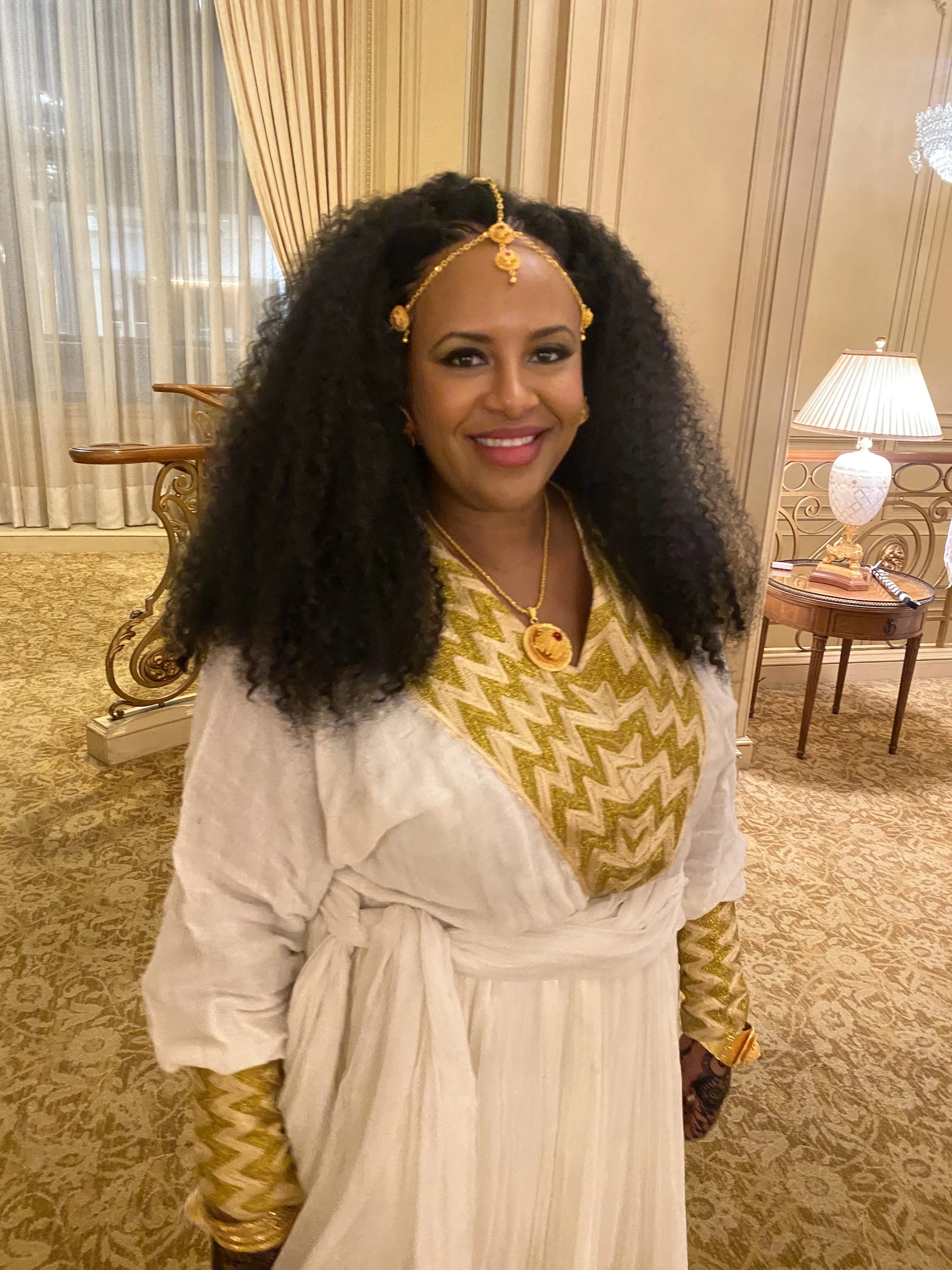 A woman dressed in traditional Indian attire, wearing gold jewelry, a white shawl, and a patterned dress, smiling in a warmly lit room with elegant furnishings and curtains.