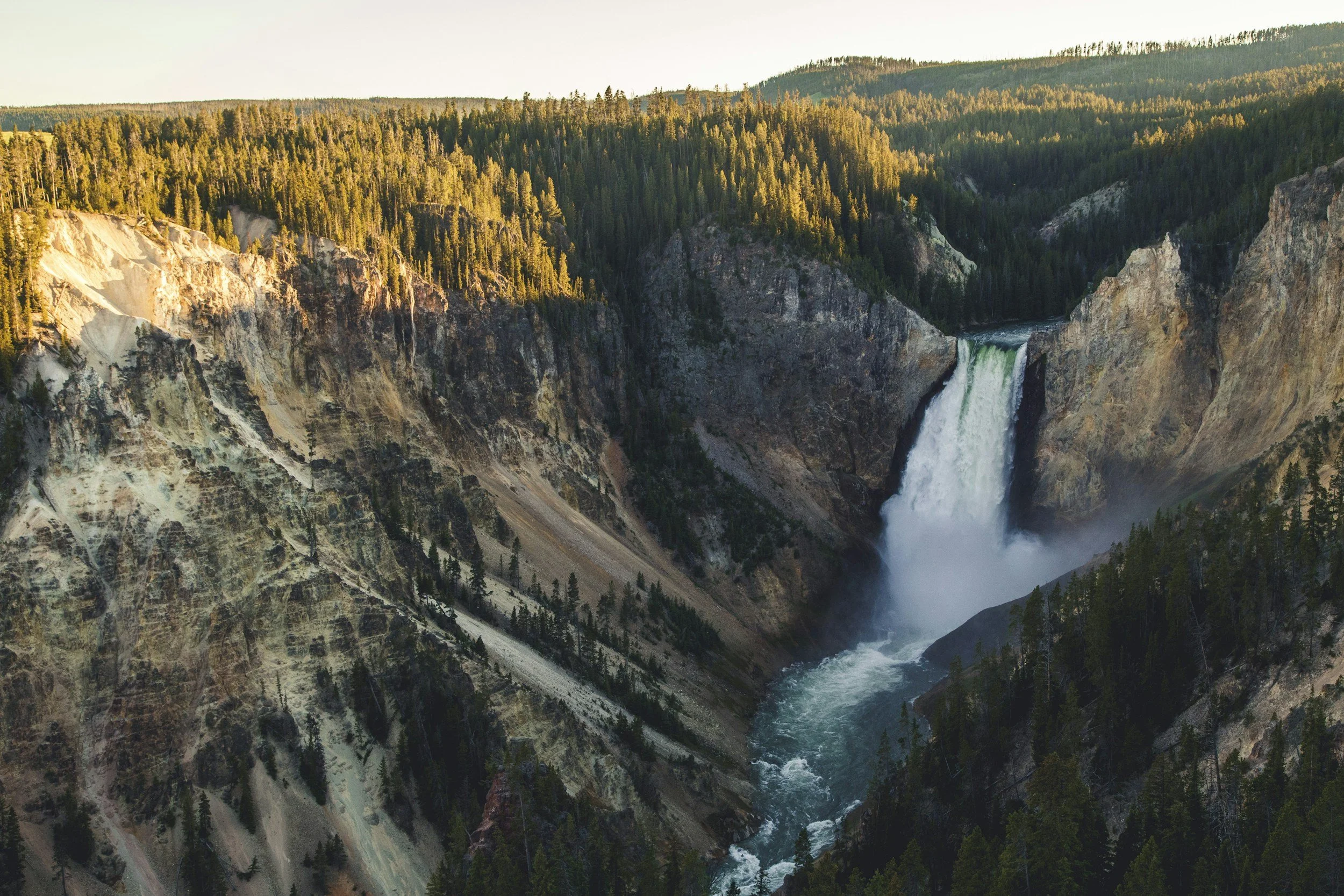 A scenic view of a waterfall cascading into a river in a canyon surrounded by trees and rugged cliffs during sunset.