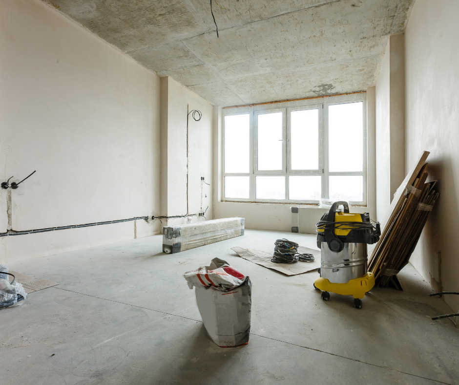 An unfinished room under construction with bare concrete floors and walls, large window letting in natural light, construction tools and materials scattered around.