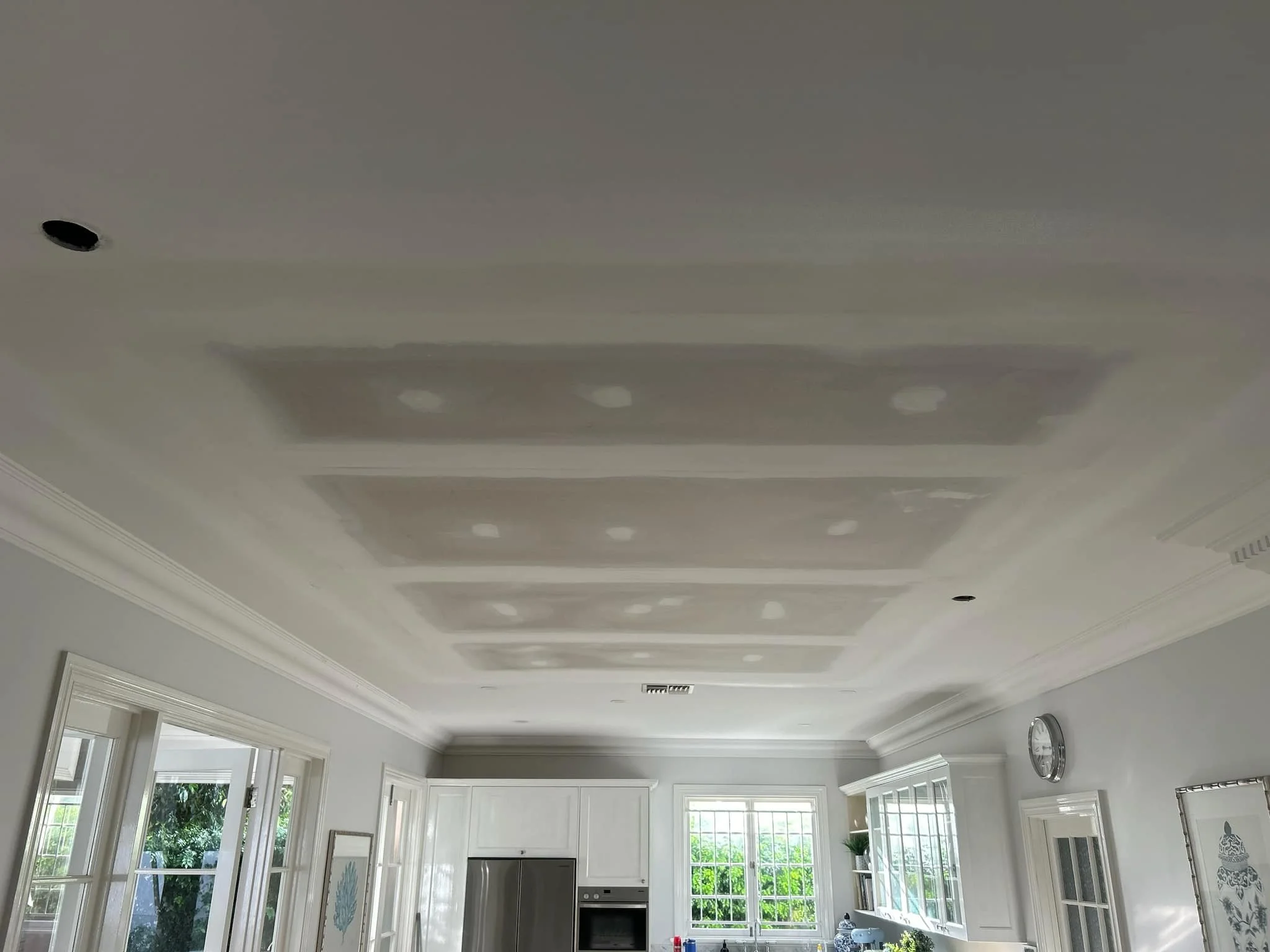 Ceiling with drywall patches and recessed lighting in a bright kitchen with white cabinets and large windows.