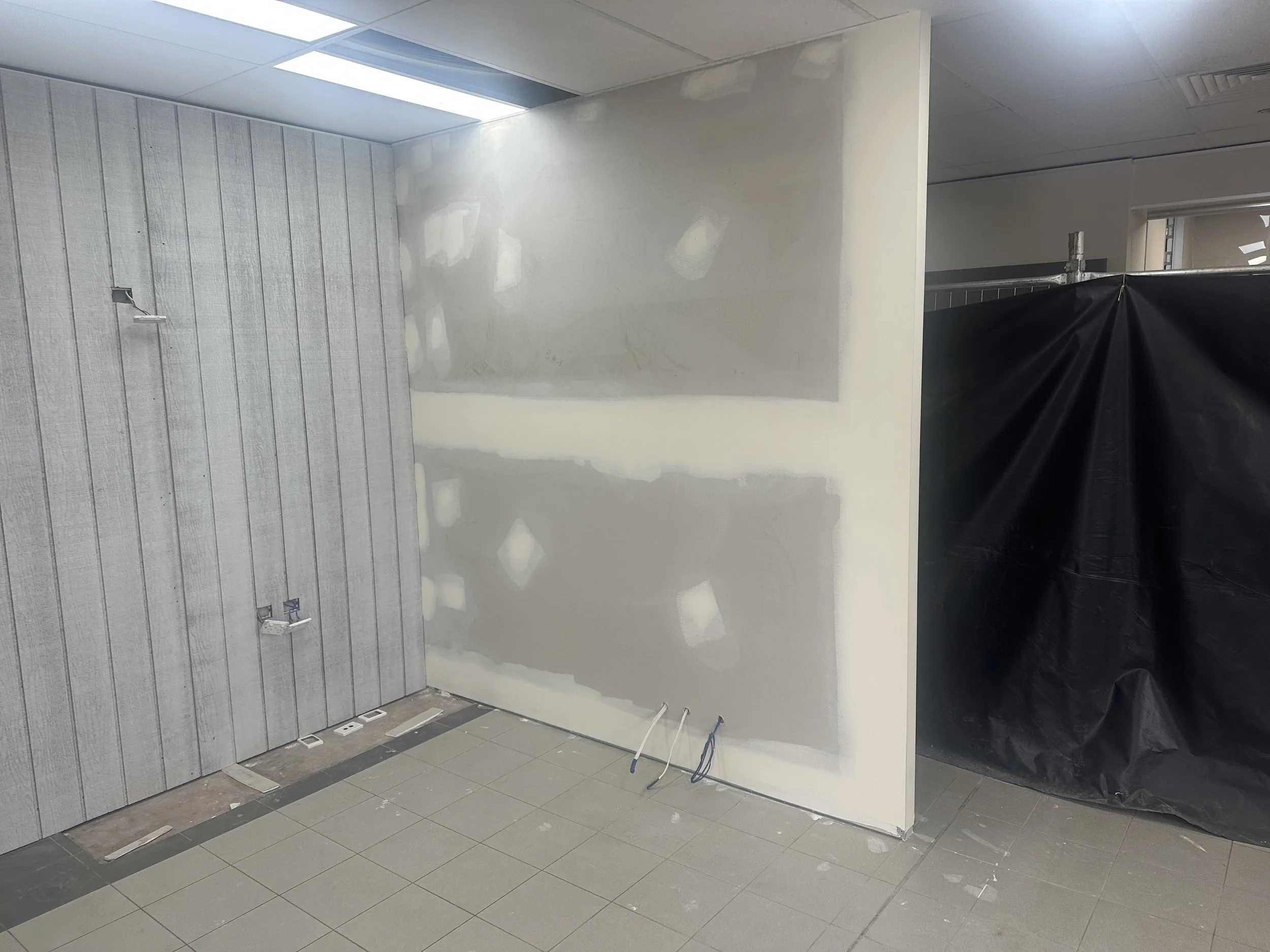 Office or commercial space under renovation with partially drywall and wall panels, construction materials, electrical wiring, and a black plastic covering.