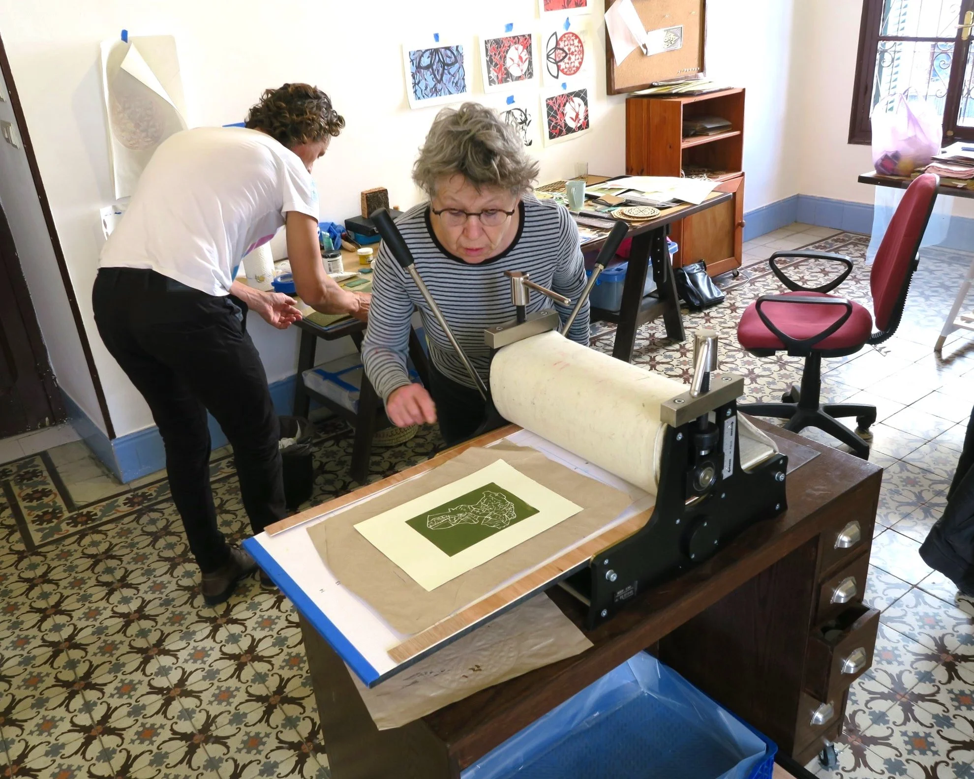 Two women working on a printmaking project in a colorful art studio, with artwork on the walls and printing supplies on the table.