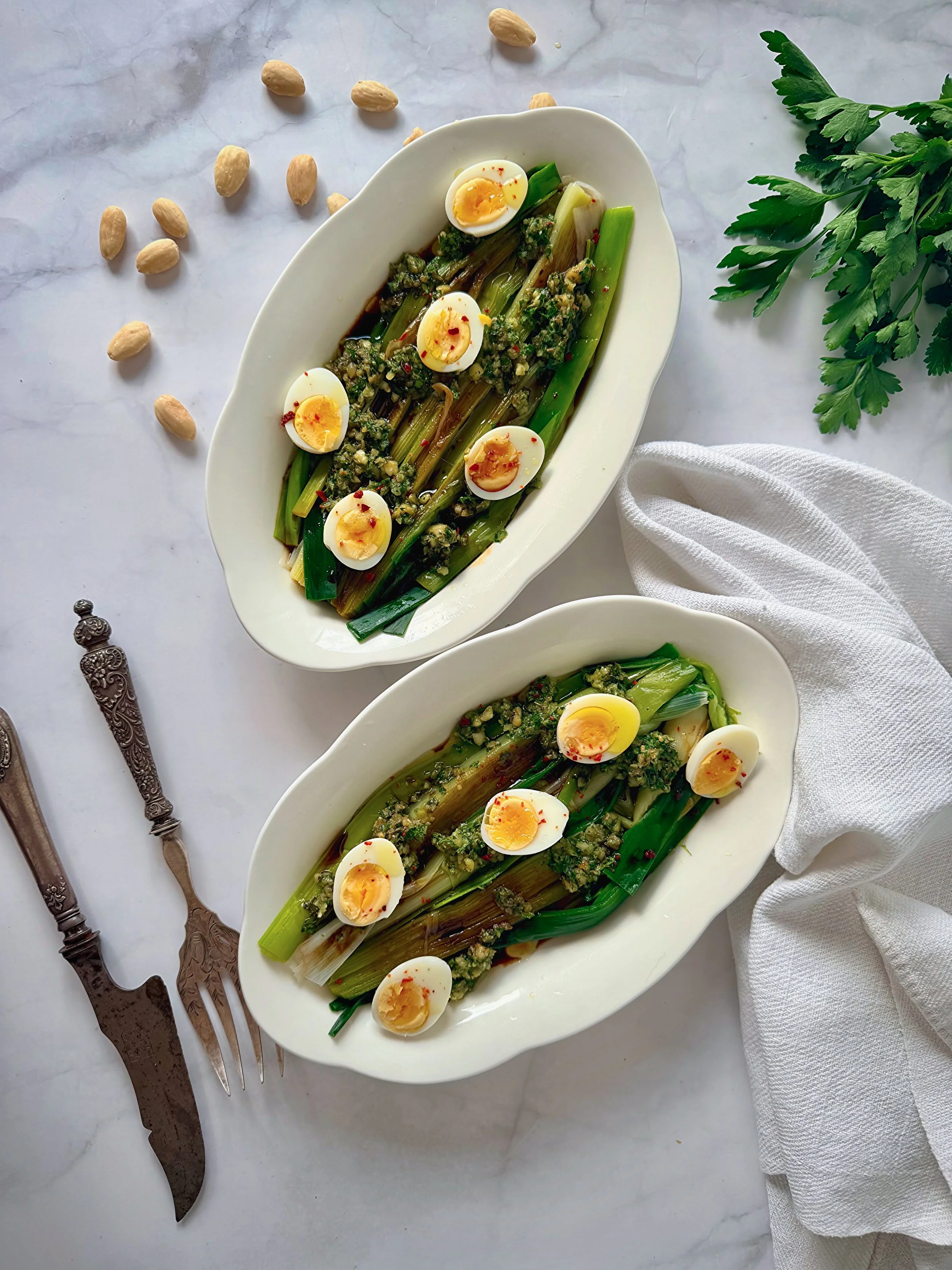 Deux plats de poireaux fondants servis avec des œufs de caille, nappés d'une réduction de vinaigre de Xérès au miel et garnis d'un condiment d'amandes torréfiées, câpres et persil, photographiés sur un fond en marbre avec des couverts anciens.