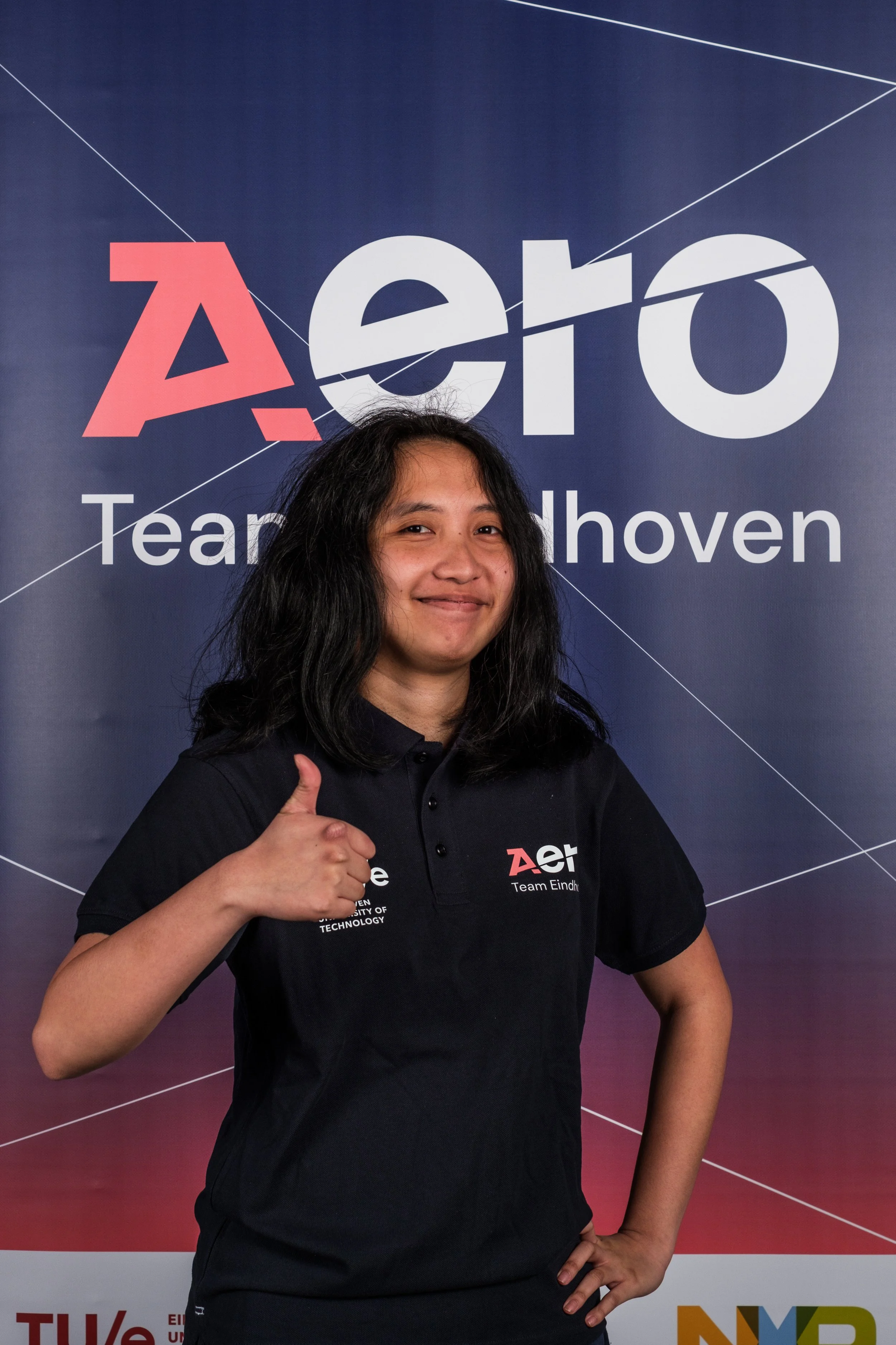 Woman with shoulder-length black hair smiling and giving a thumbs up in front of a blue backdrop with the text 'Aero Team Eindhoven' and the logo.