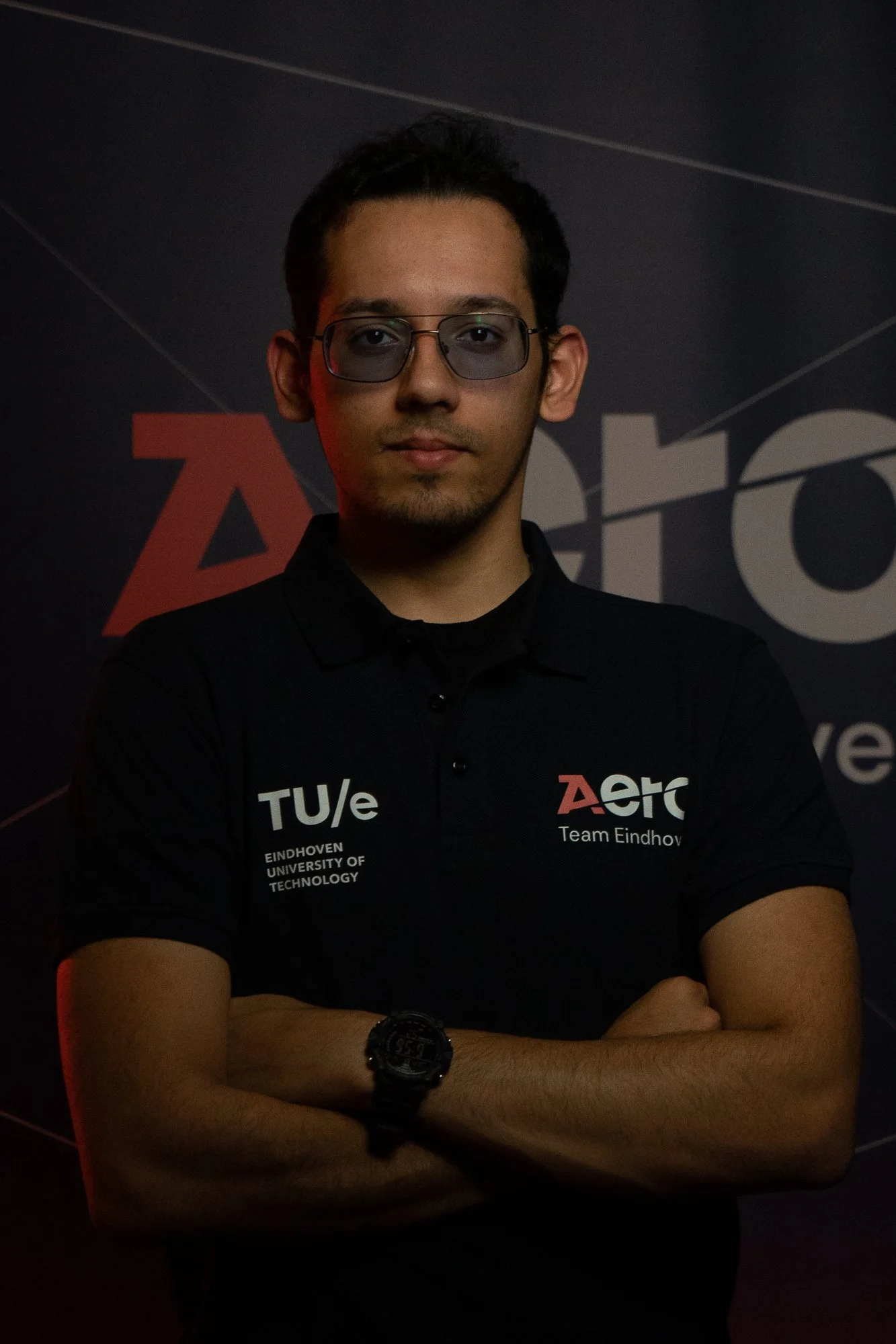 Young man with dark hair and glasses, wearing a black polo shirt with logos and text, standing with arms crossed against a dark background with partially visible logo and text.
