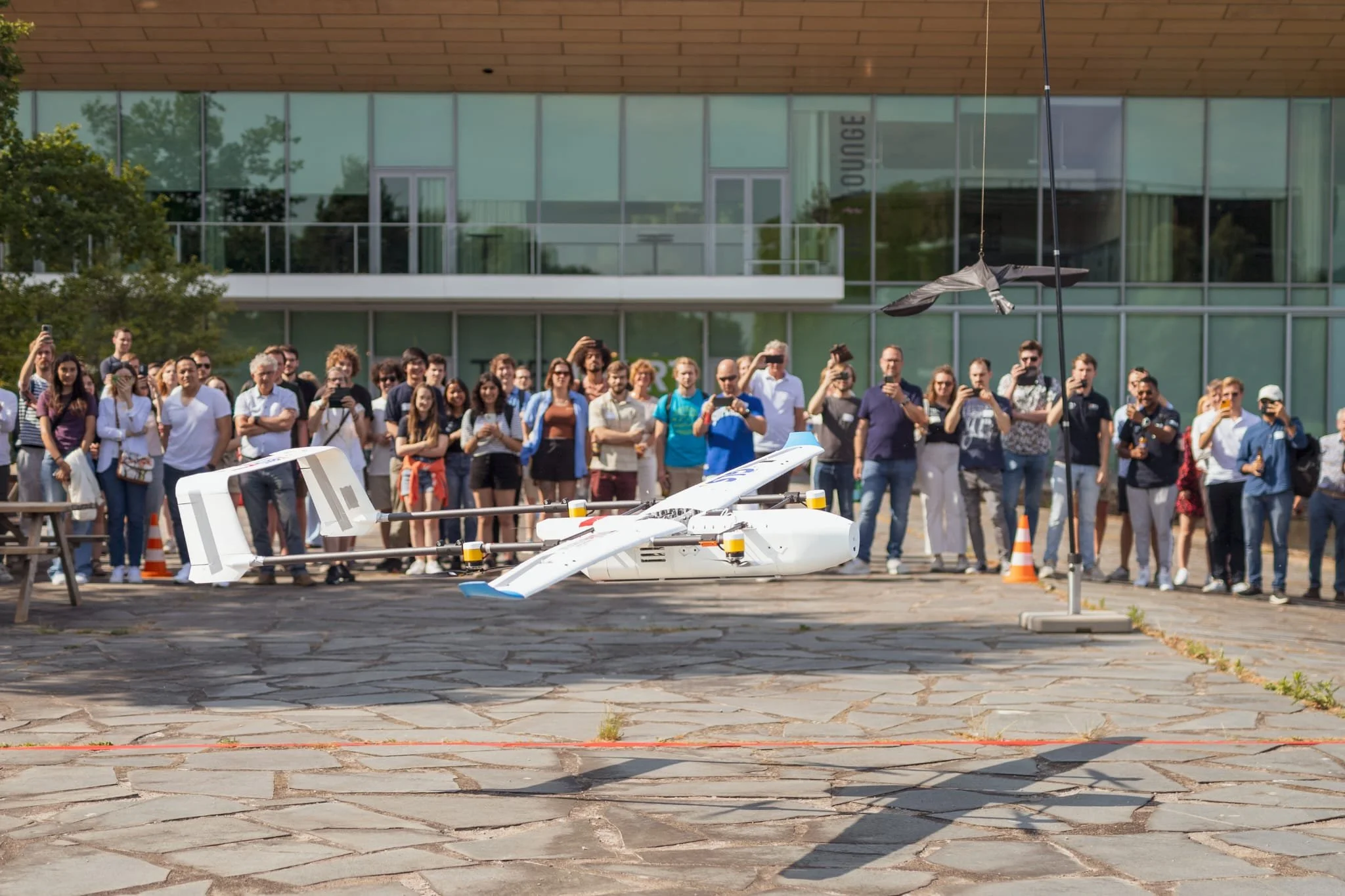 A group of people watching a large drone flying close to a suspended object outdoors in front of a modern building.