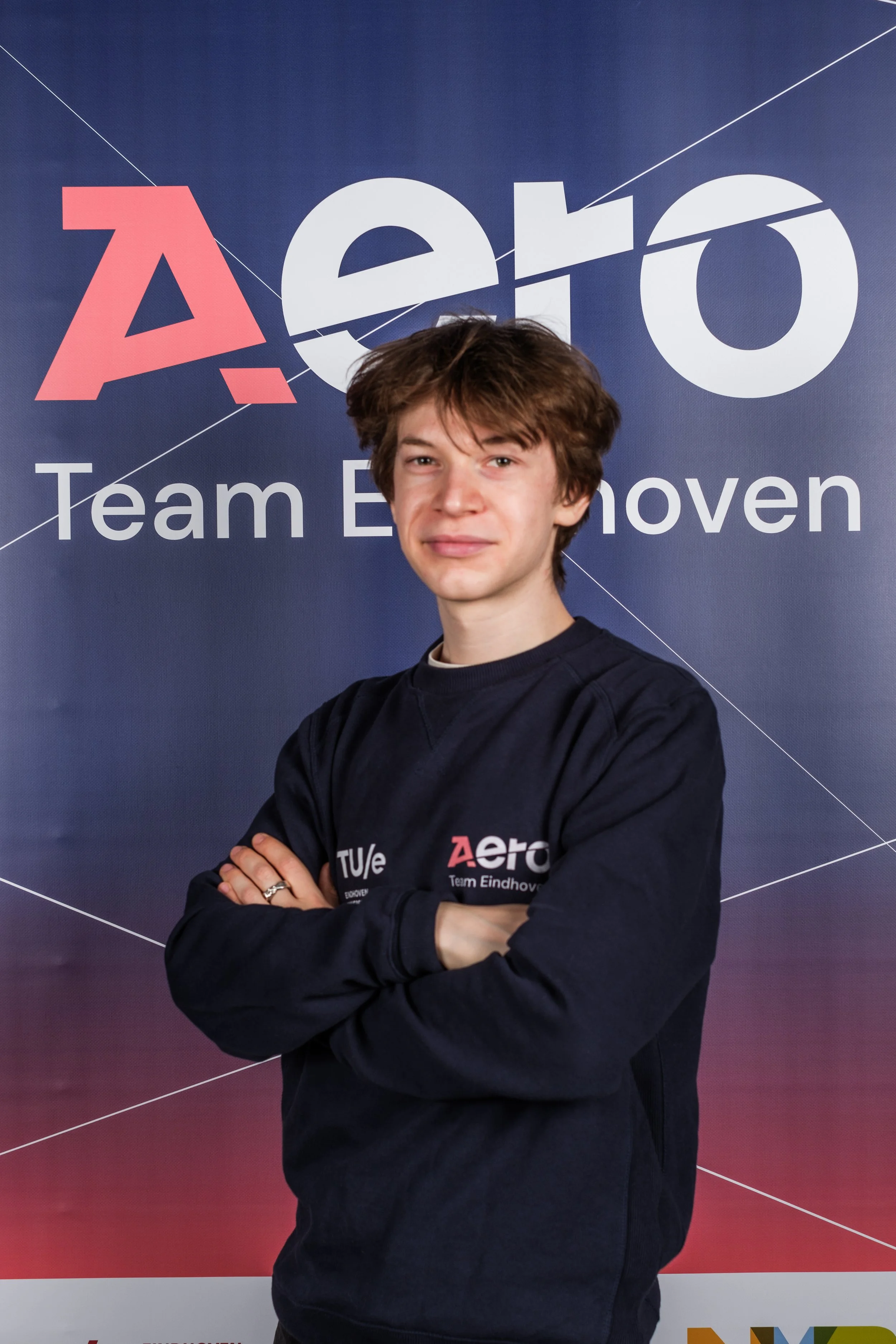 Young man with brown hair standing with arms crossed, wearing a black Aero team sweatshirt, in front of a blue background with the Aero logo and text that reads "Team Eindhoven."