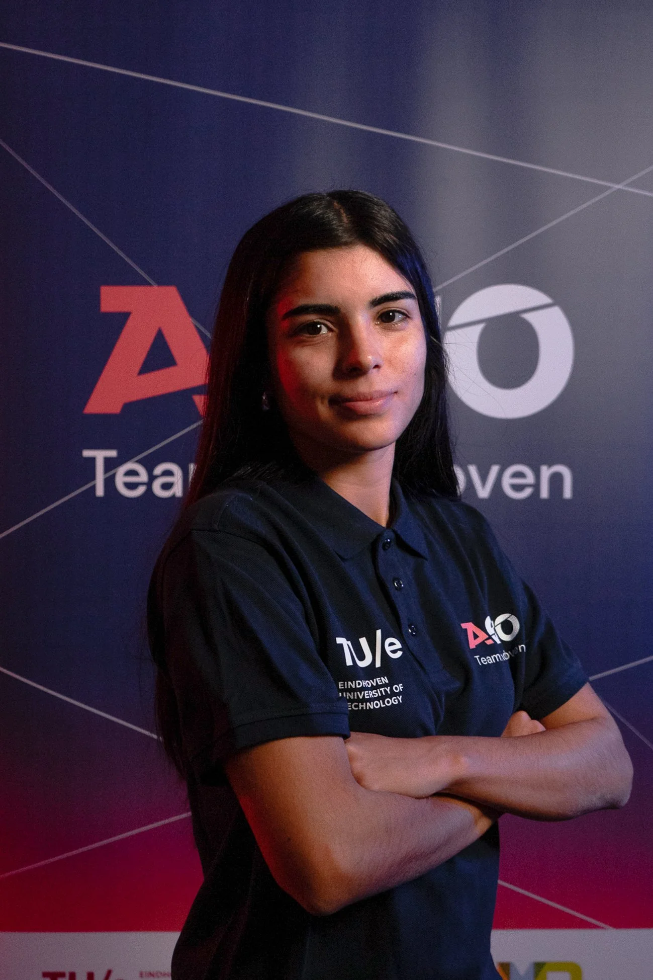 A woman with long dark hair and crossed arms standing in front of a dark blue background featuring logos for Team Eindhoven, TU/e, and Eindhoven University of Technology.