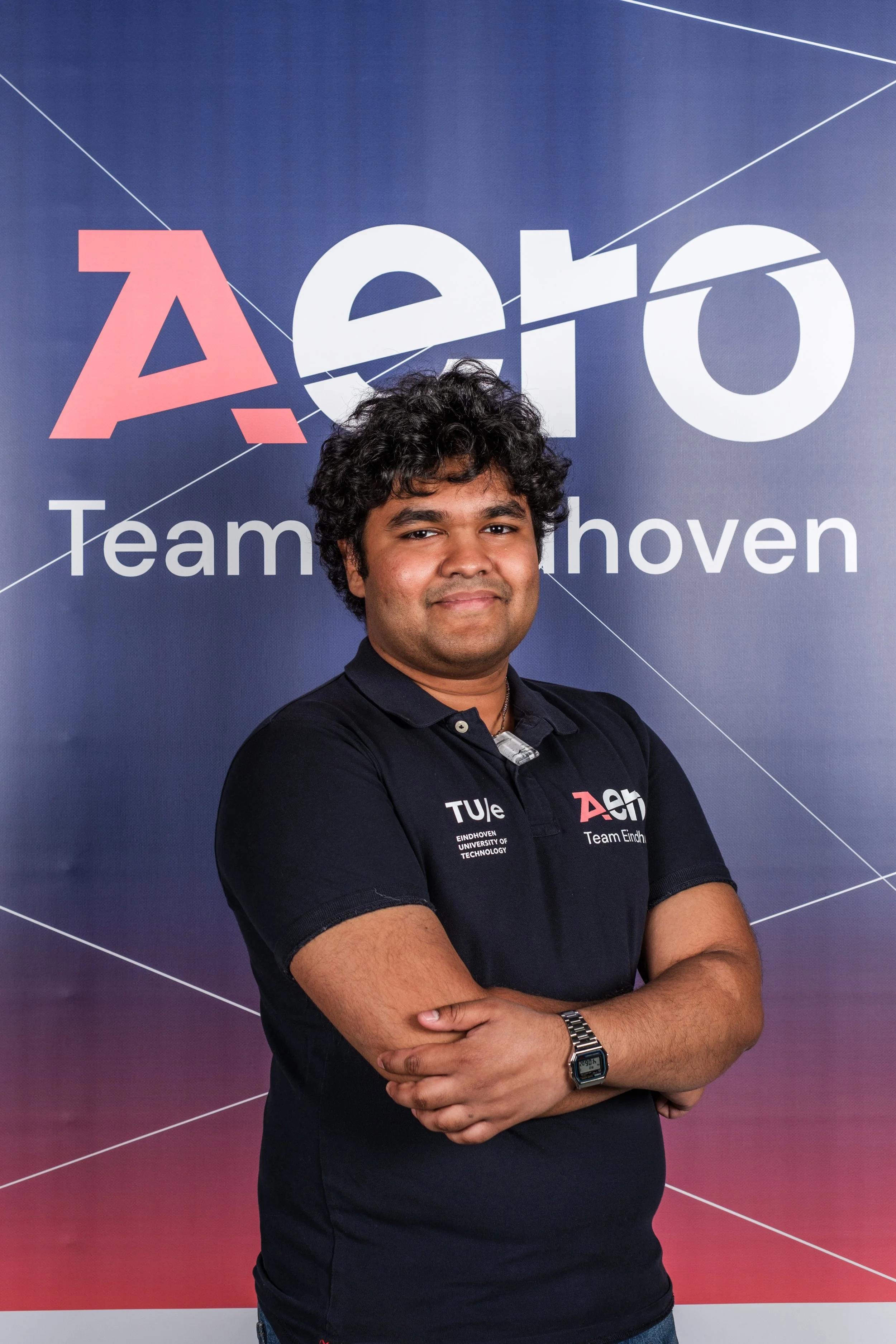 A man with curly black hair wearing a black polo shirt with the logos of Eindhoven University of Technology and Team Eindhoven, standing in front of a large backdrop with the words "EHTO" and "Team Eindhoven."