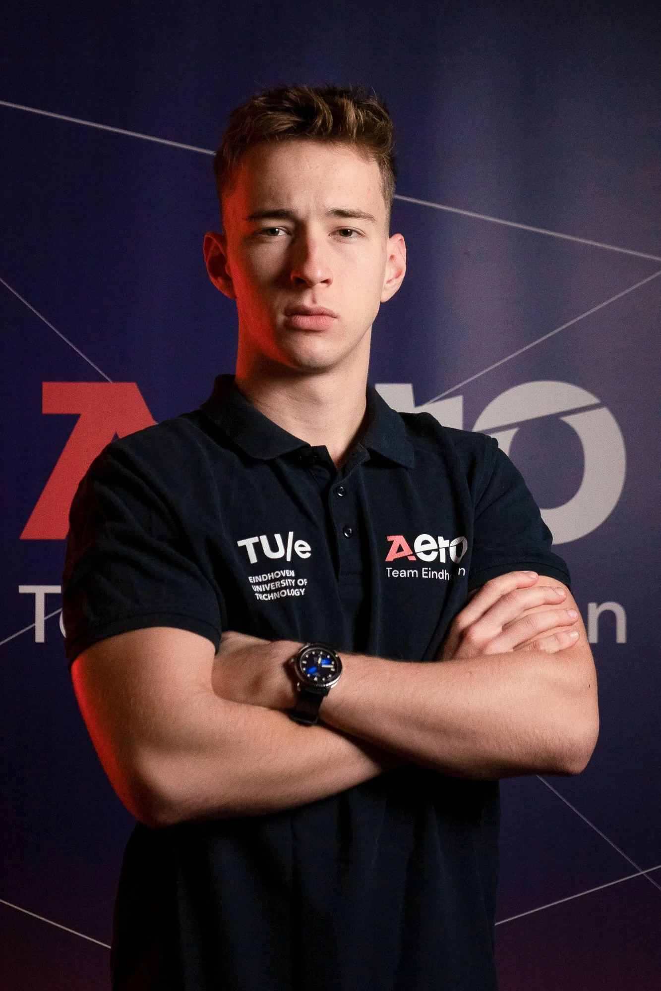 Young man with crossed arms wearing a black polo shirt with logos, standing in front of a dark background with text and graphical elements.