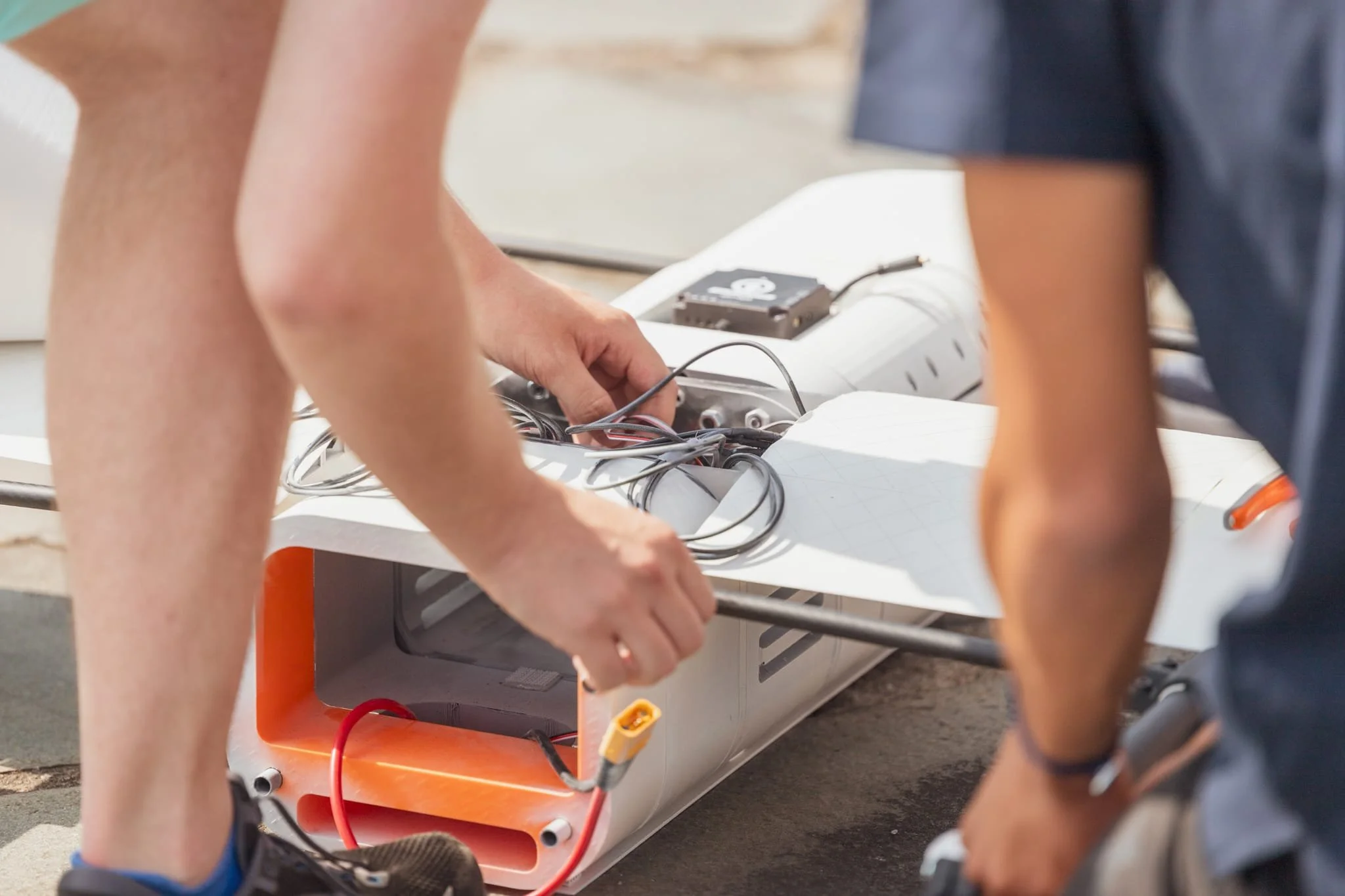 Two people work on the electronics of a small, white, skateboard-like device outdoors. One person is reaching into the device, handling wires, while the other person watches. The device has a red wire and visible internal components.