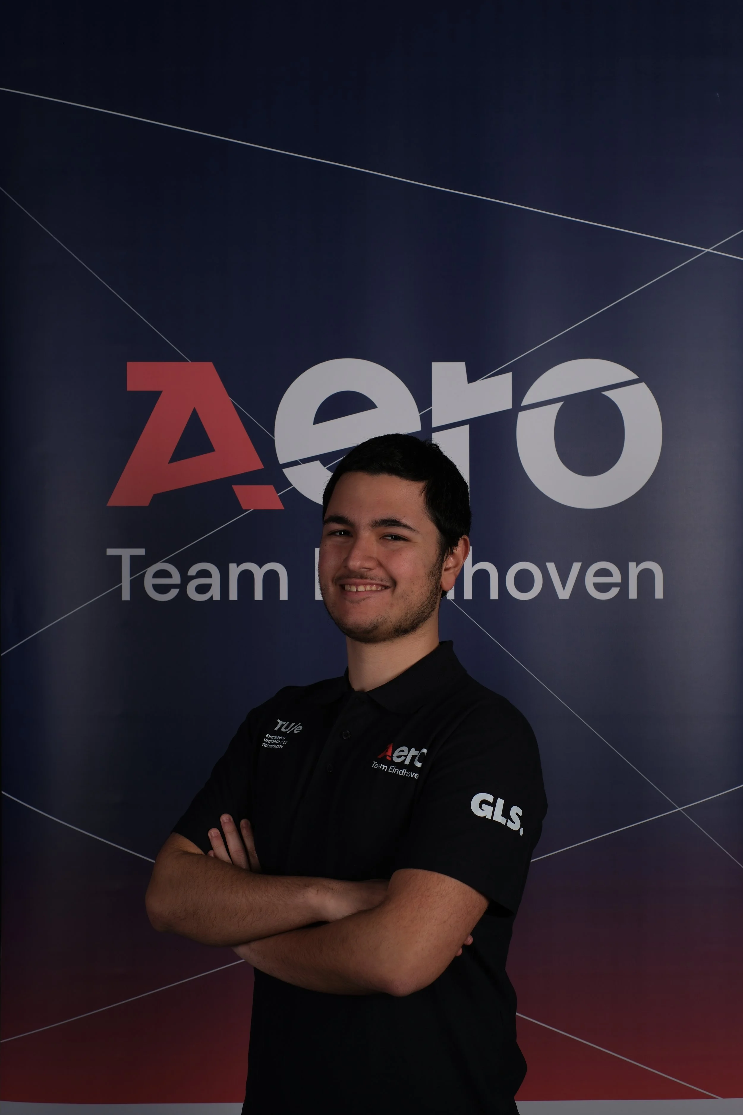 A young man smiling with arms crossed, wearing a black polo shirt with logos, standing in front of a backdrop with the text 'Aero Team Eindhoven'.