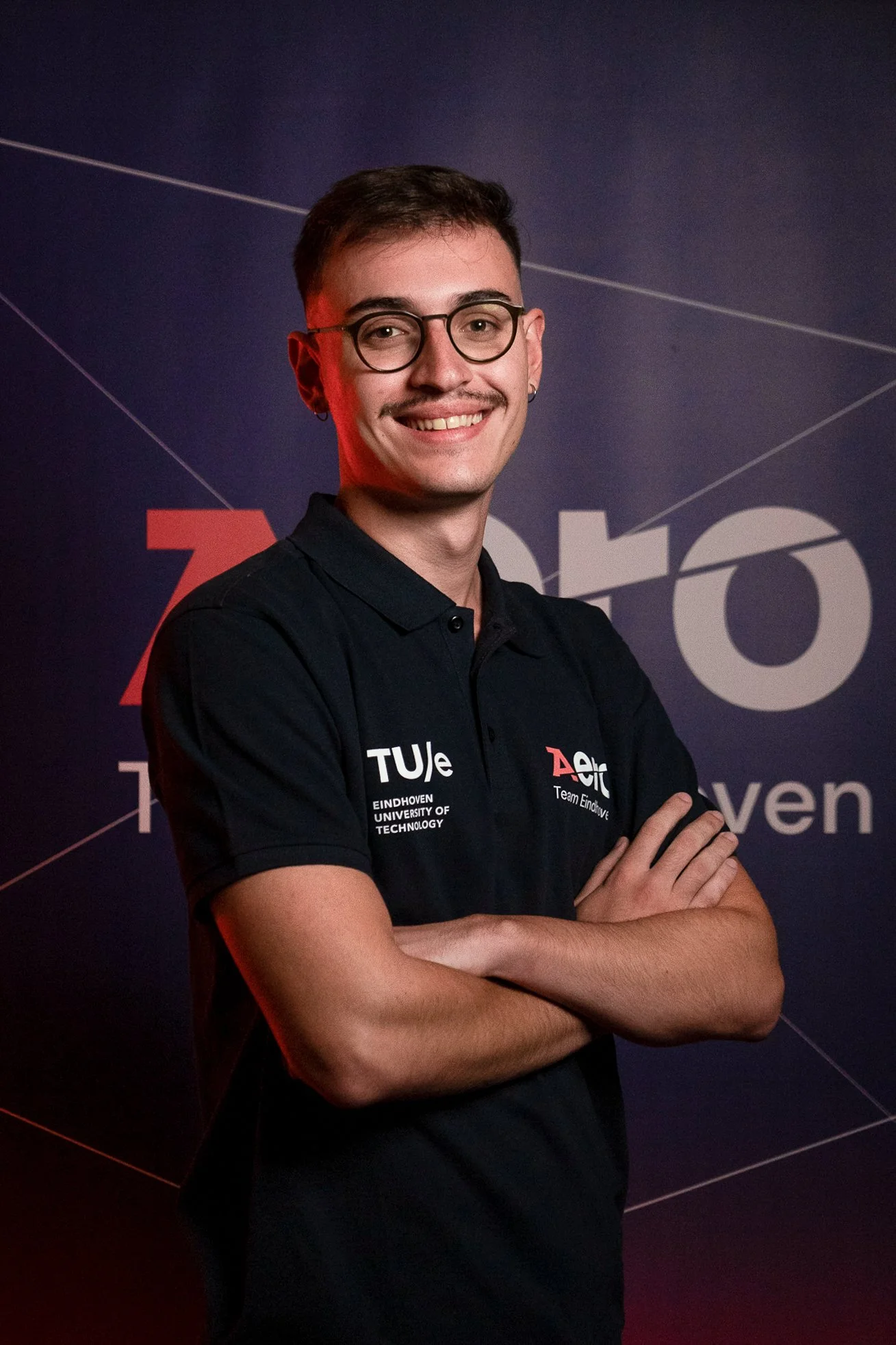 Young man with glasses, smiling, wearing a black polo shirt with logos, standing with arms crossed in front of a dark background with text and geometric lines.
