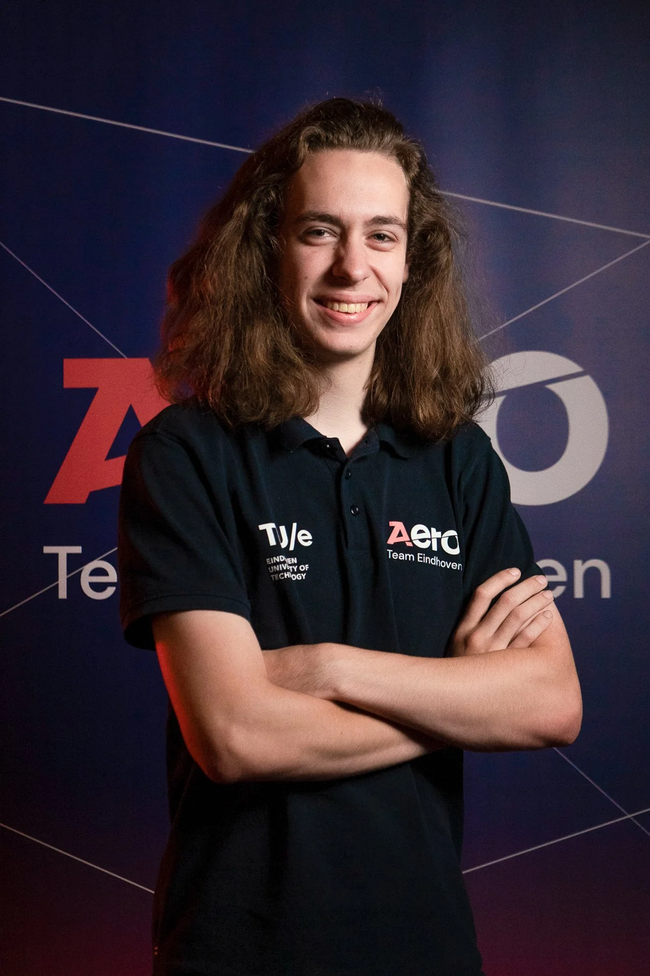 Young man with long, curly hair standing with arms crossed in front of a dark background with logos and text, wearing a navy polo shirt with logos of Eindhoven University of Technology and Team Eindhoven.