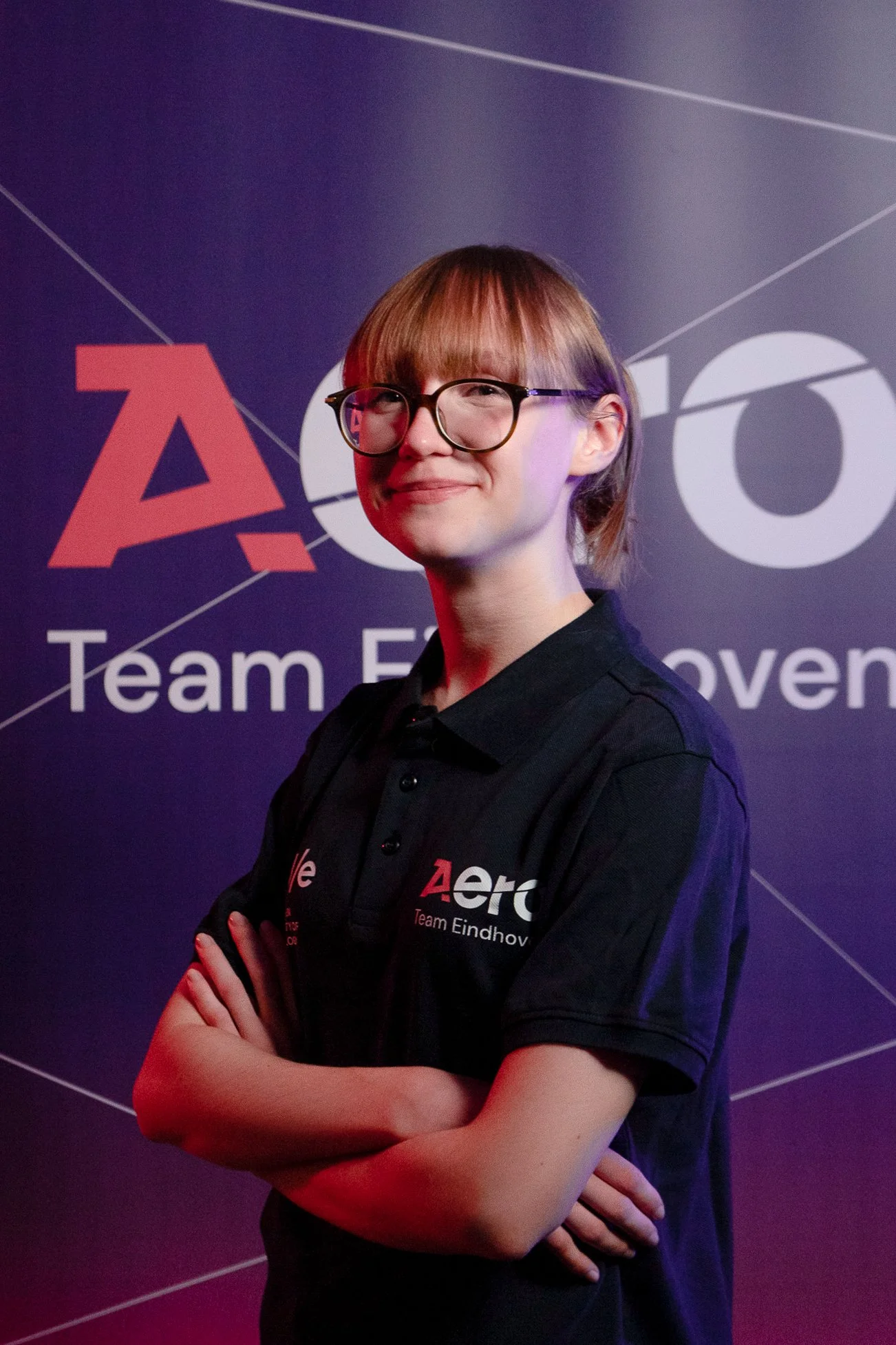 A young woman with glasses, wearing a black polo shirt with "Aero" and "Team Eindhoven" logos, standing with arms crossed in front of a dark background with the same logos.
