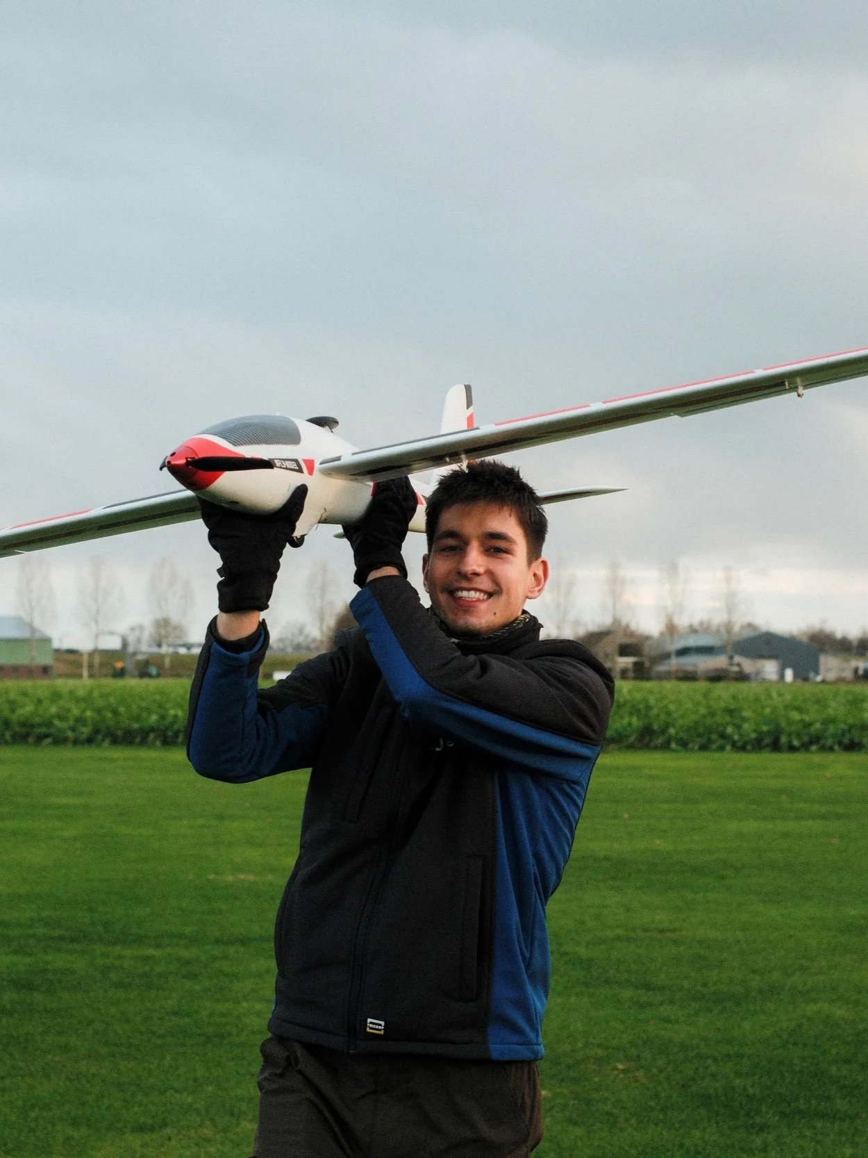 A young man smiling outdoors, holding a model airplane above his head, standing on grassy field with trees and buildings in the background.