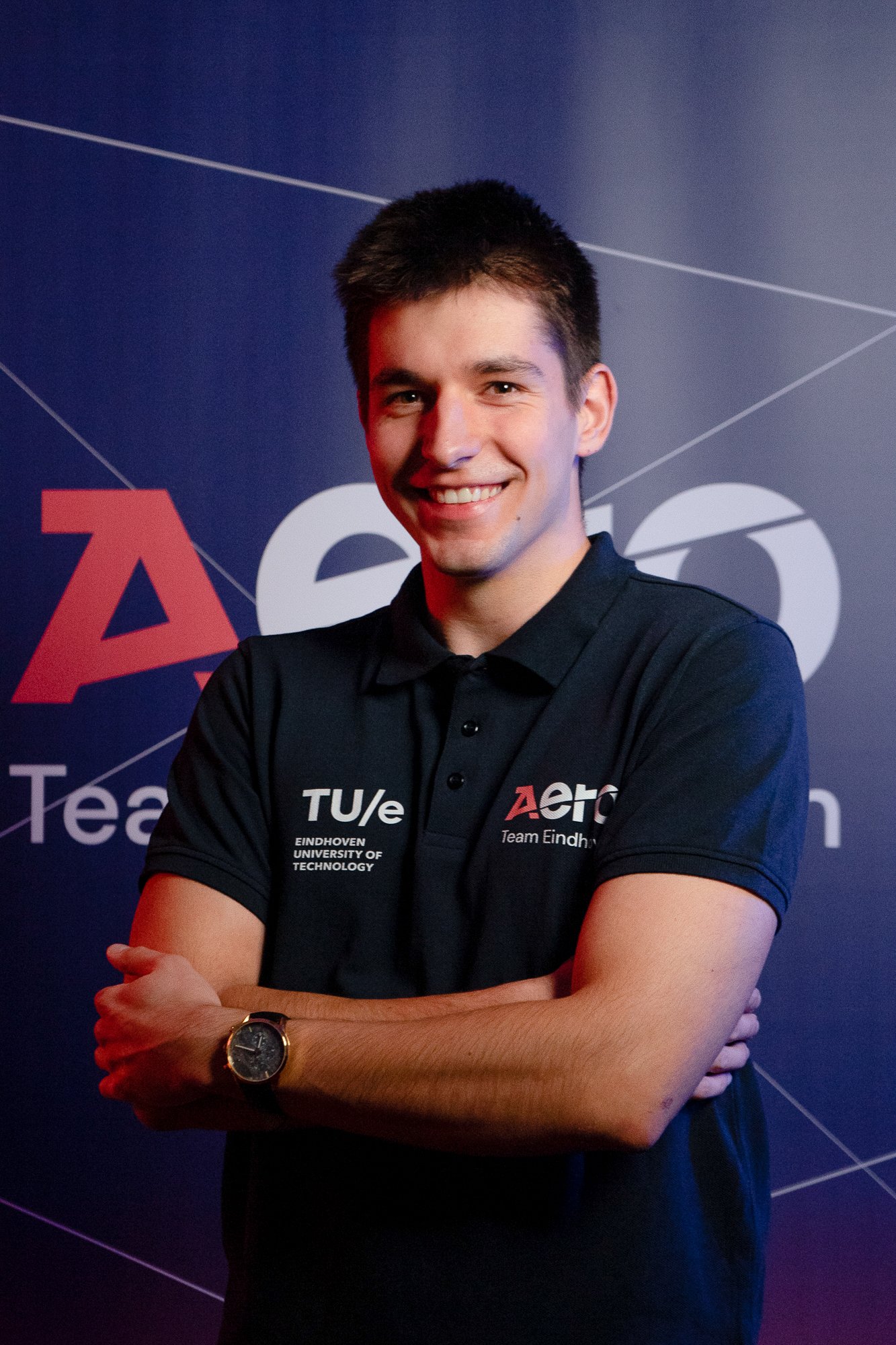 A young man smiling, standing with arms crossed, in front of a dark background with logos and text, wearing a black polo shirt with white and red text.