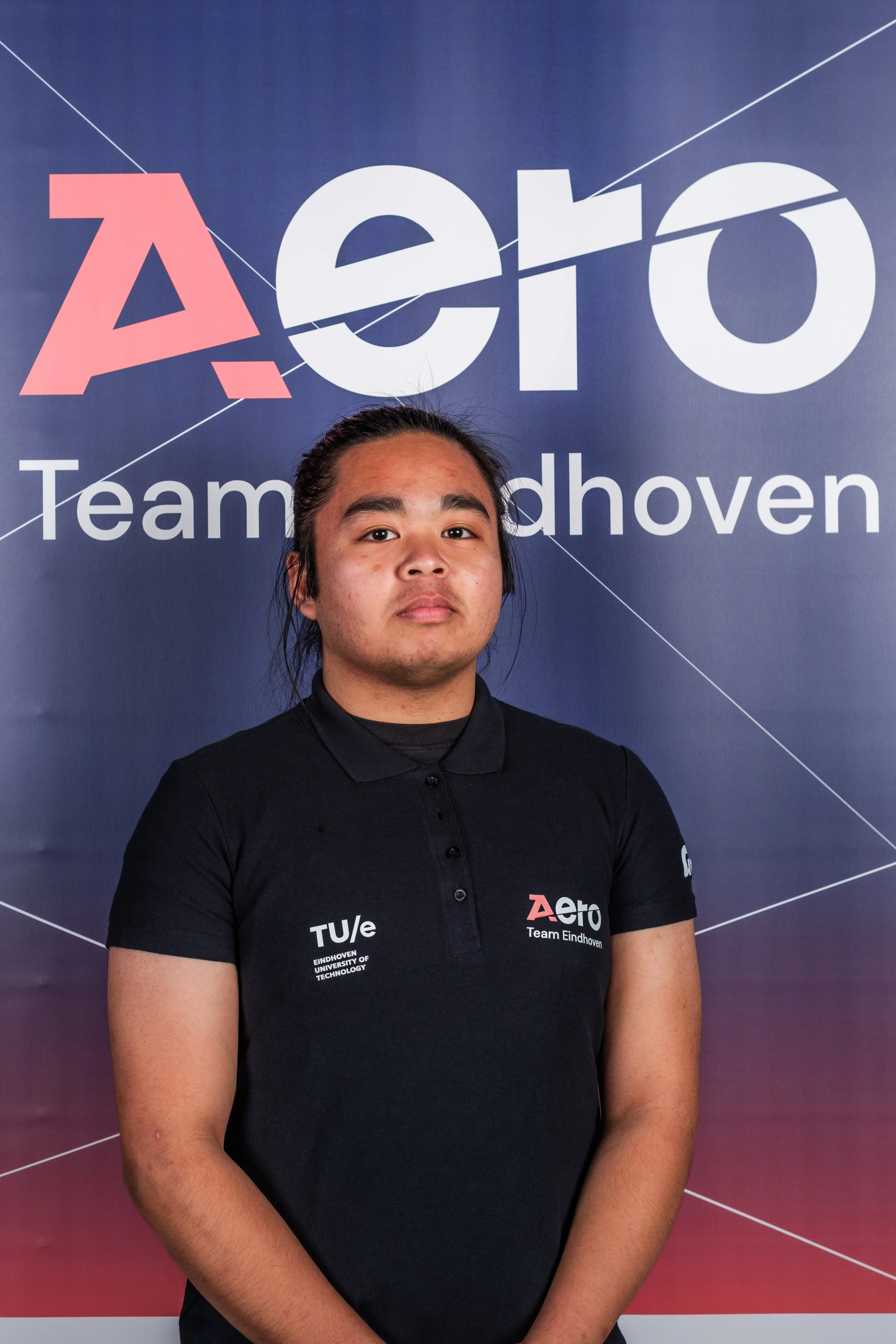 A young man with long dark hair tied back, wearing a black polo shirt with logos and text, stands in front of a blue background with the words 'Aero Team Eindhoven' printed on it.