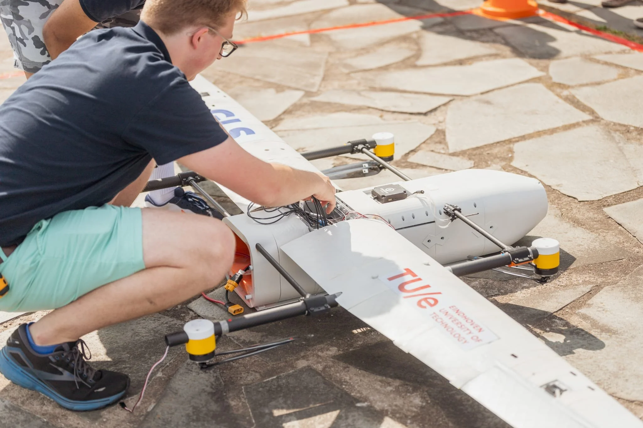 Person working on a white drone labeled "Tuße" on a stone surface, with wires and electronic components visible.