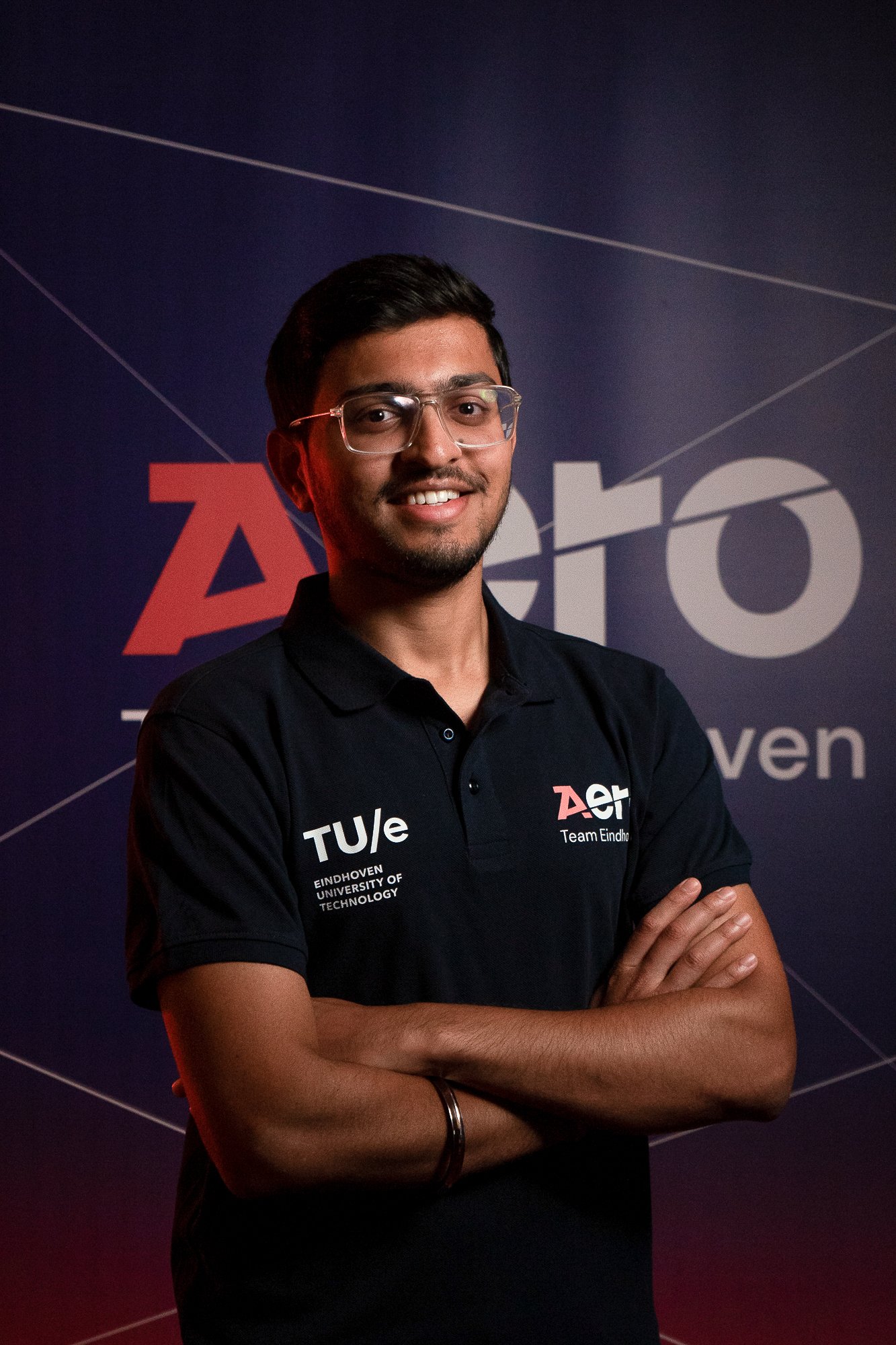 A man standing in front of a backdrop with 'Aero' and 'TUE' logos, wearing a black polo shirt with university logos, smiling and crossing his arms.