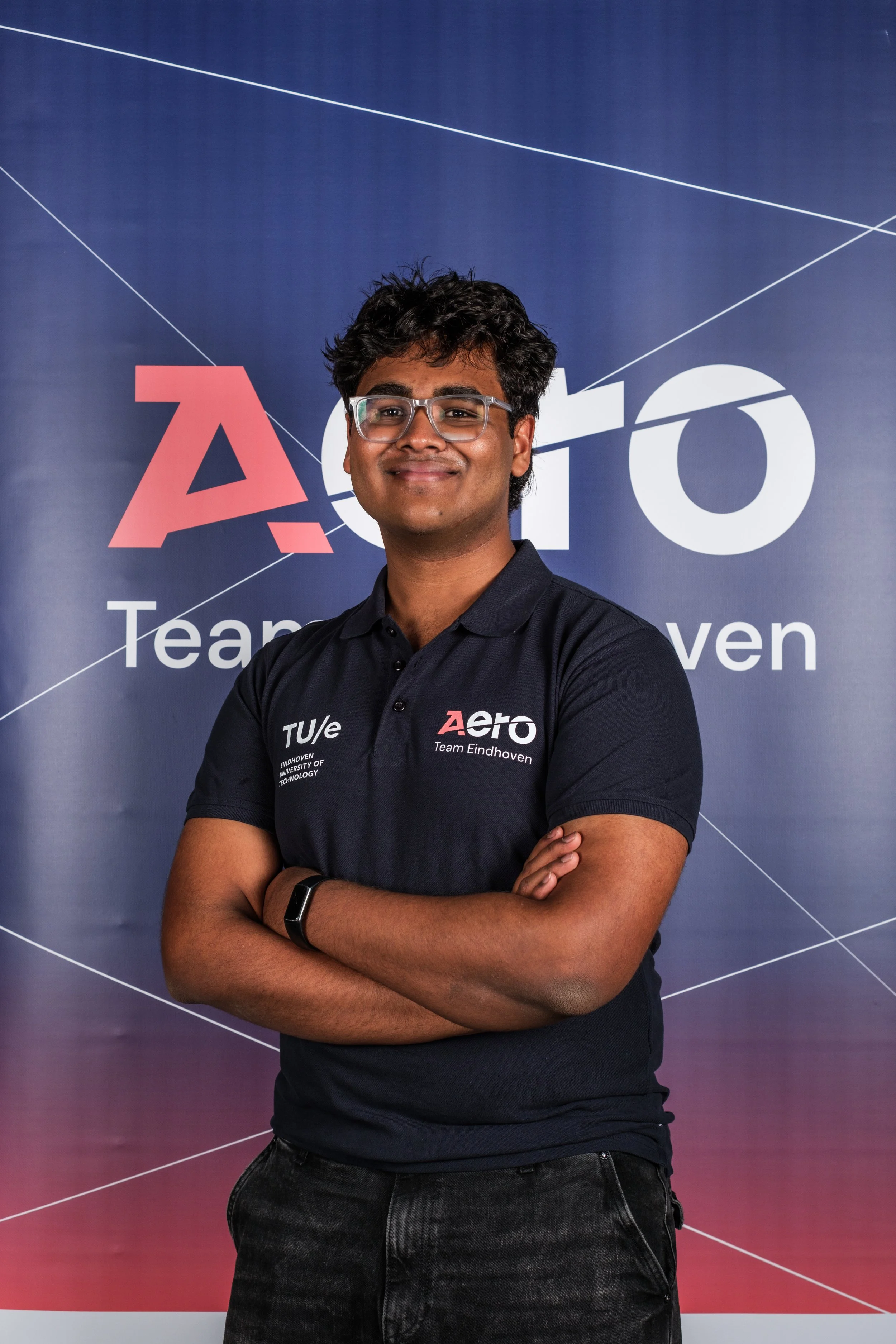 A young man standing with arms crossed, smiling, in front of a blue backdrop with 'Aero' and 'Team Eindhoven' logos.