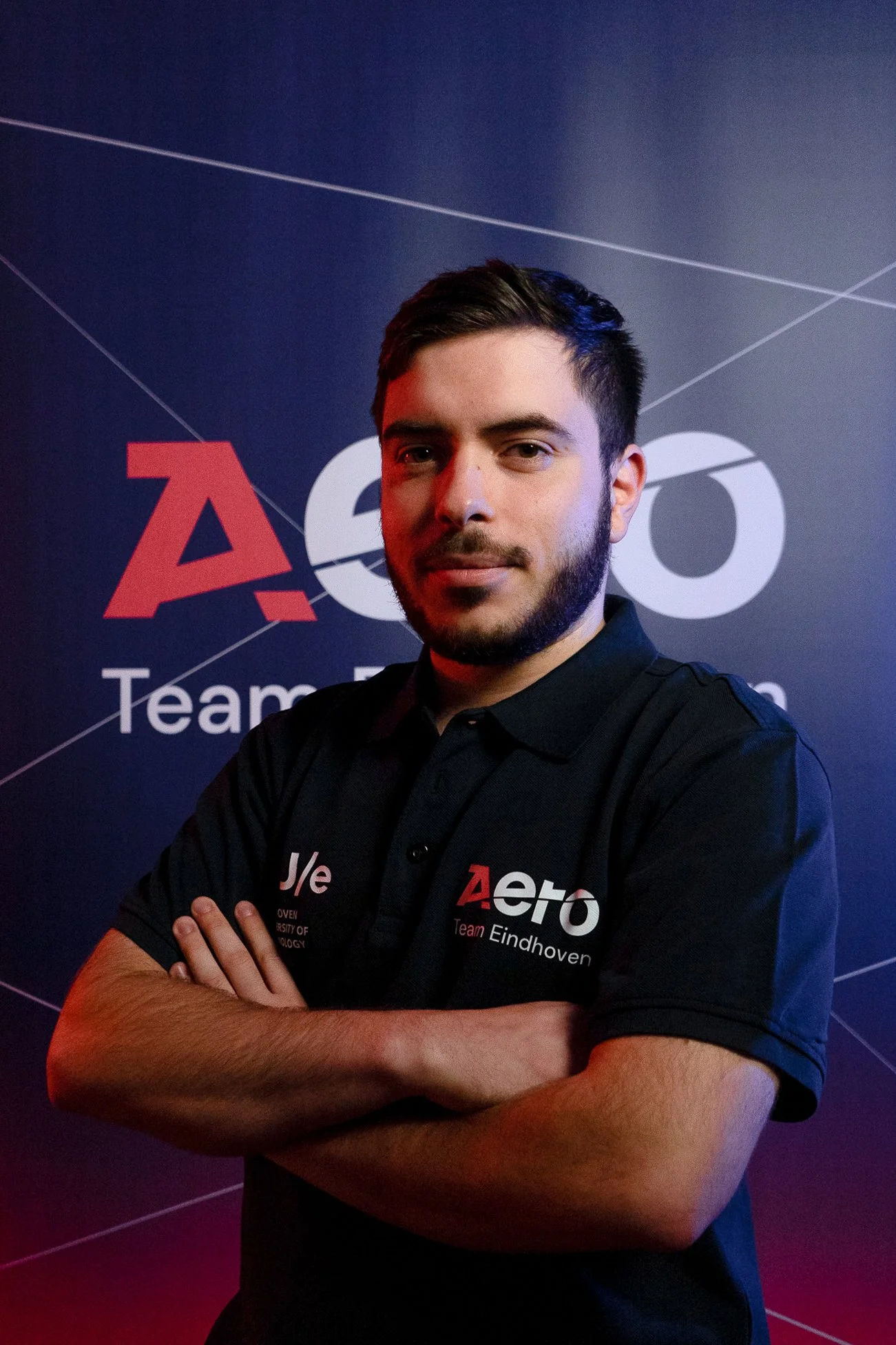 A man with dark hair and a beard, wearing a black polo shirt with logos, standing with arms crossed in front of a dark background with logos and text for 'Aero Team Eindhoven'.