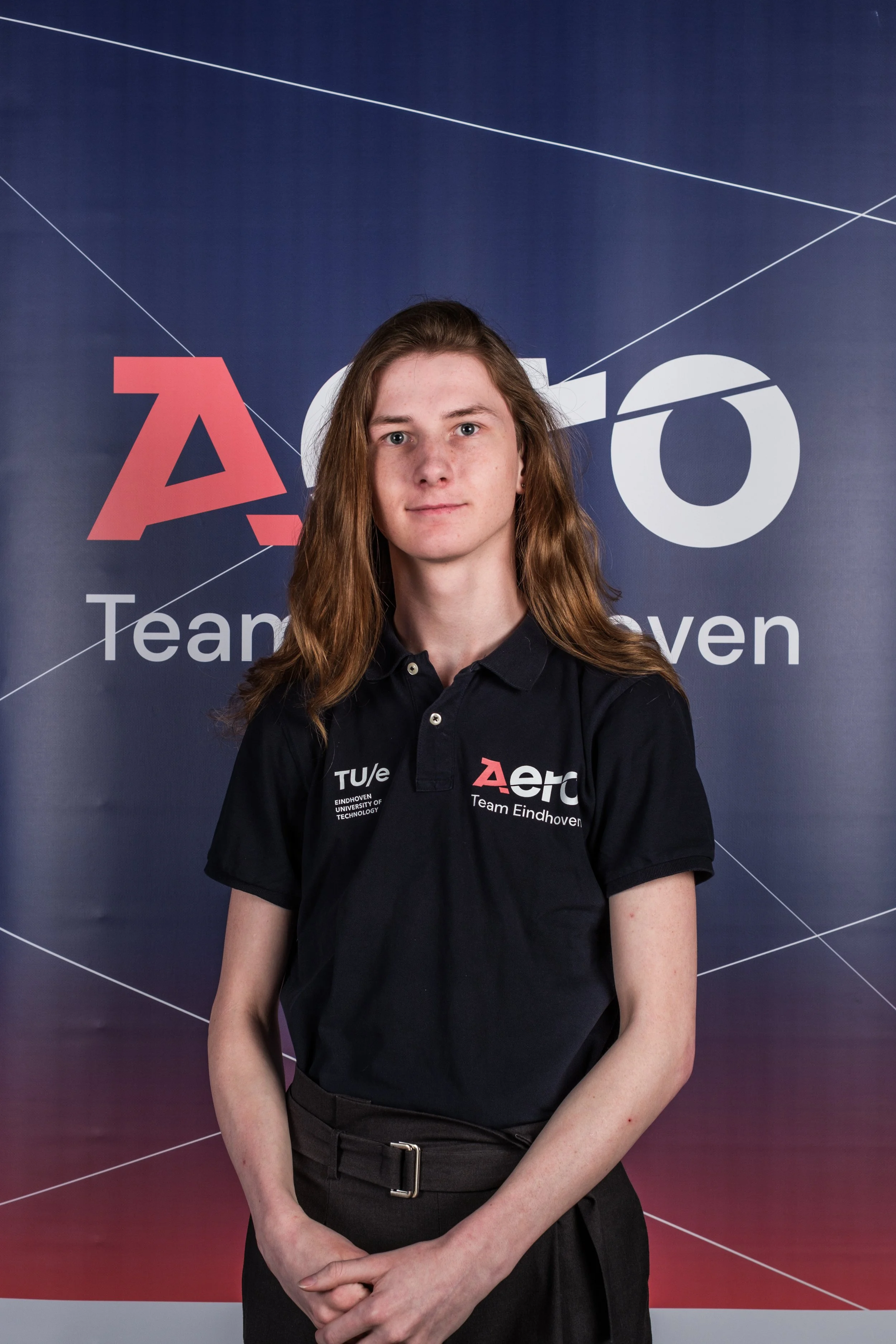 Young woman with long red hair posing in front of a backdrop with the 'Aero' and 'Team Eindhoven' logos. She is wearing a black polo shirt with similar logos.