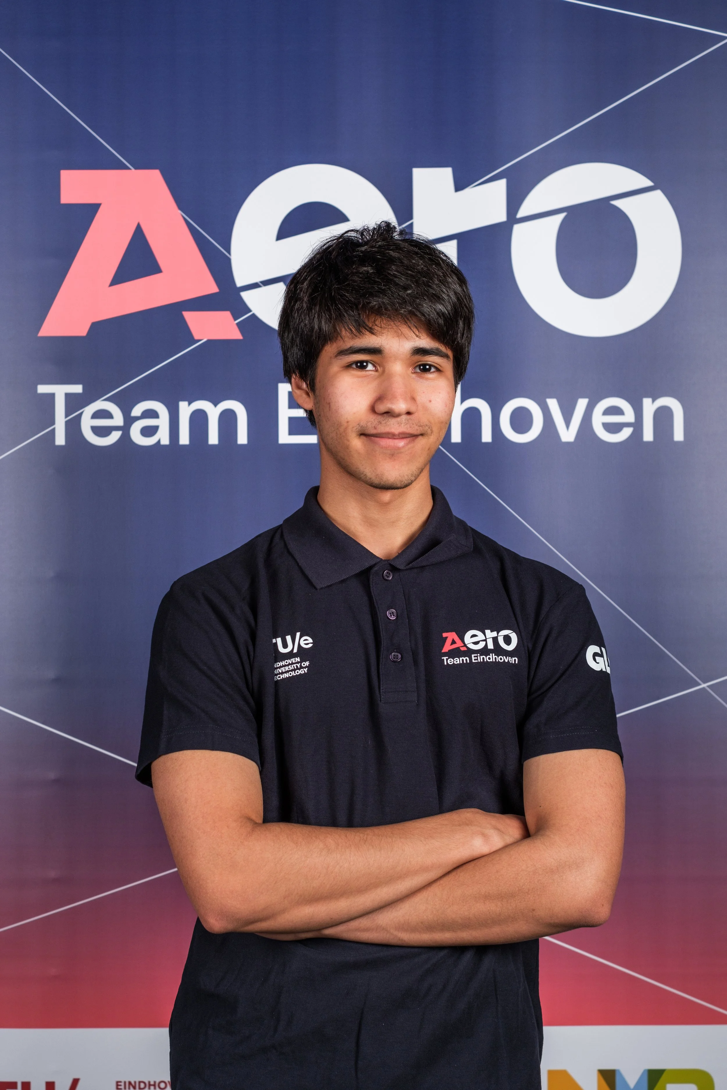 Young man with dark hair, wearing a navy polo shirt with logos, standing cross-armed in front of a blue backdrop with 'Aero Team Eindhoven' text.