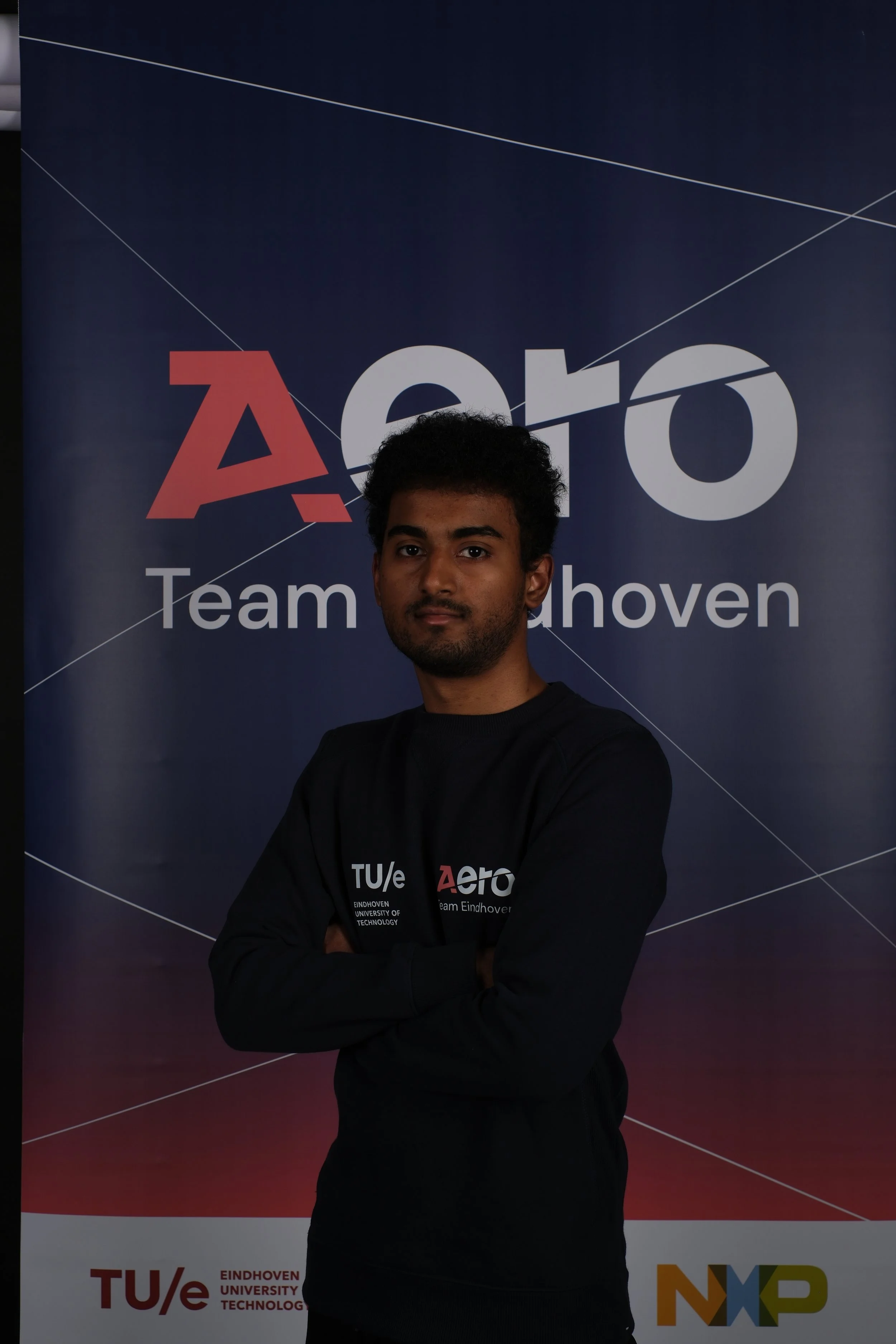 A young man with dark curly hair and a beard standing with arms crossed in front of a large banner that reads 'Aero Team Eindhoven' and displays the logos of TU/e Eindhoven University of Technology and NXP.