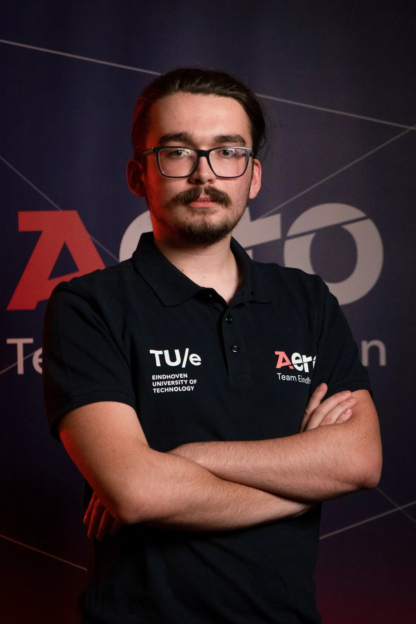 Young man with glasses, mustache, and beard wearing a black polo shirt with logos, standing with arms crossed in front of a dark promotional backdrop.