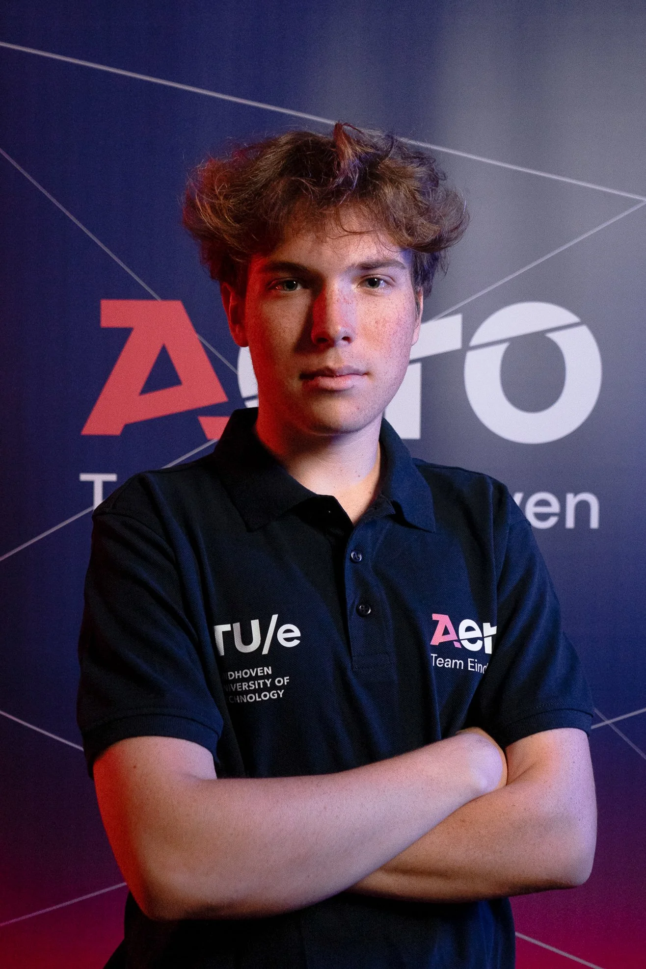 A young man with curly hair standing with arms crossed in front of a backdrop with the 'auto' and 'Team Eindhoven' logos, wearing a black polo shirt with logos on the chest.