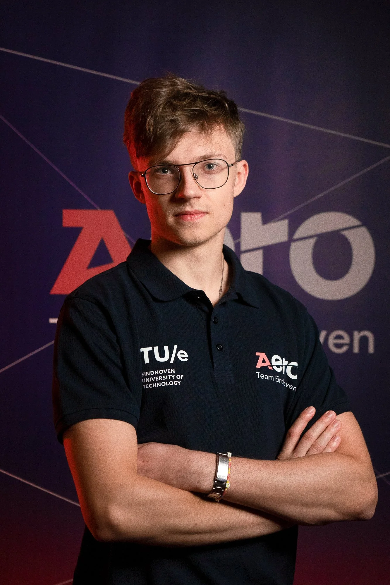 A young man with glasses and short brown hair, wearing a black polo shirt with logos and text on it, standing with arms crossed against a dark background with geometric lines and partial text.