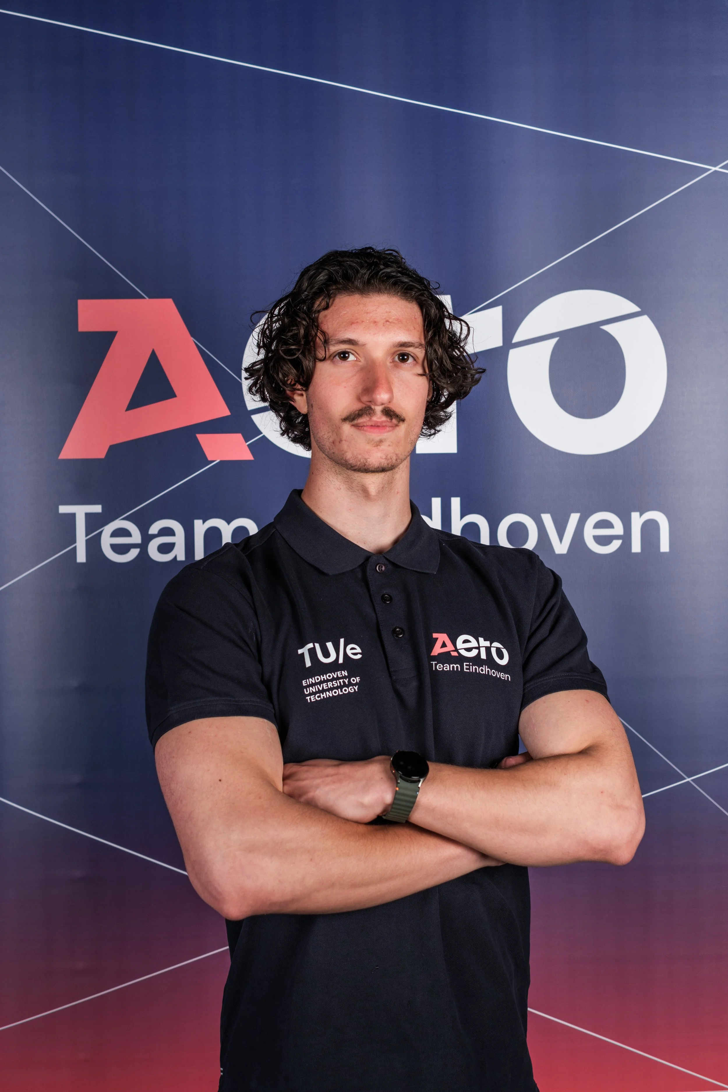 Young man with curly hair and mustache standing with arms crossed in front of a backdrop with logos and text related to Eindhoven University of Technology and Team Eindhoven.