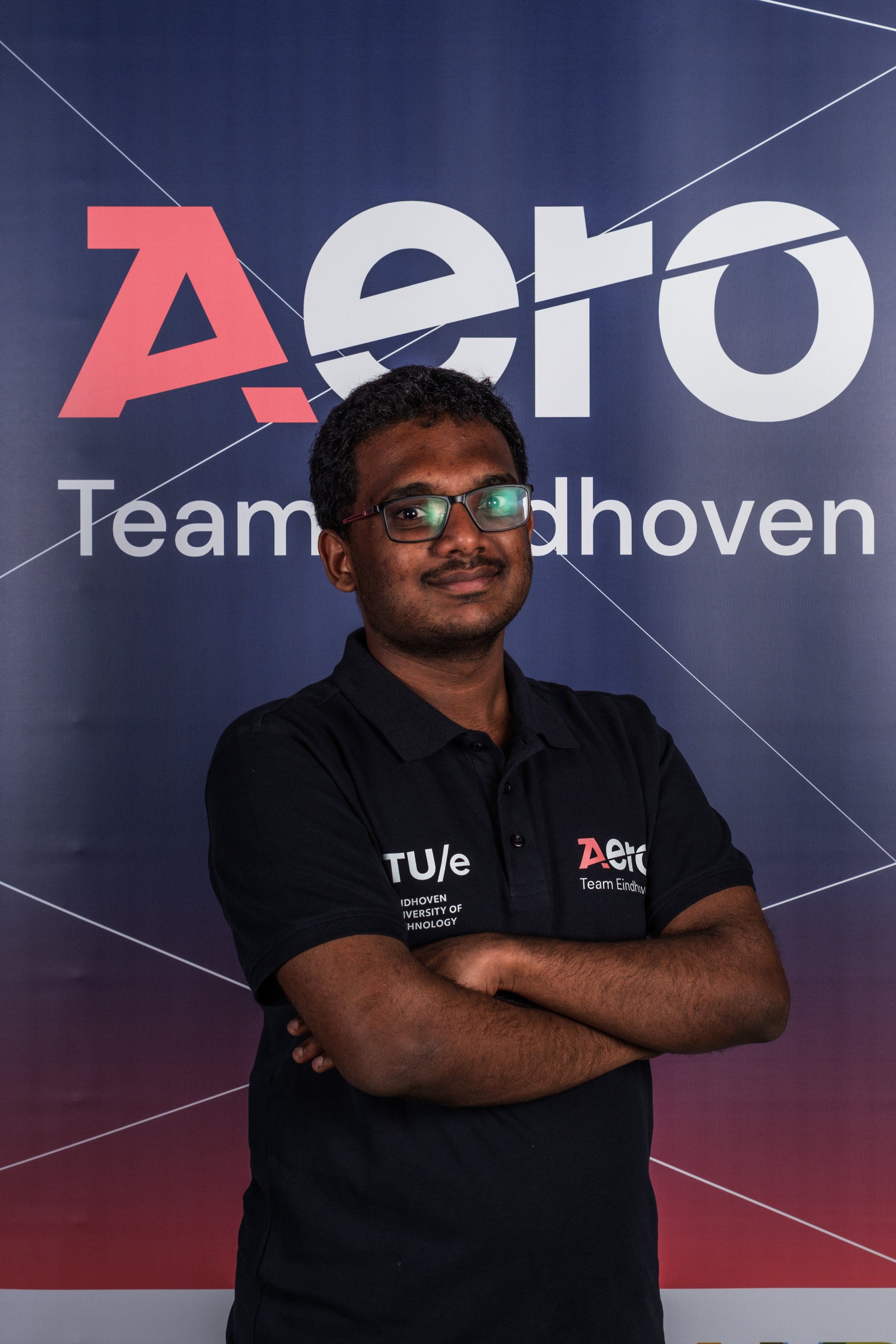 A young man with glasses standing with crossed arms in front of a dark blue background displaying the logo and text for Team Eindhoven, part of TU/e, Eindhoven University of Technology.