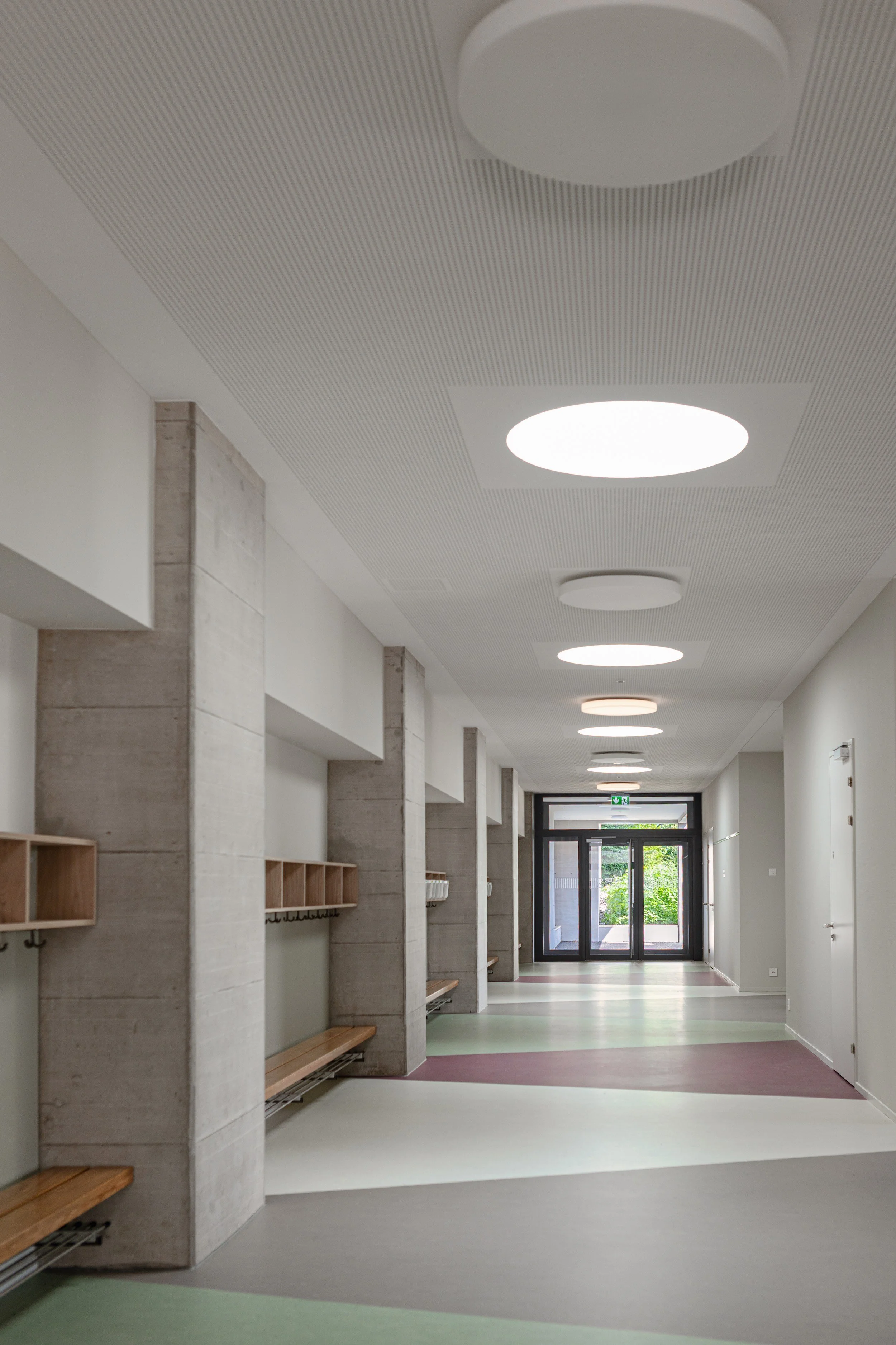 Langer Korridor im Kindergarten mit rhythmischer Lichtführung durch runde Deckenleuchten und Sichtbeton-Wänden. Starke Tiefenwirkung.