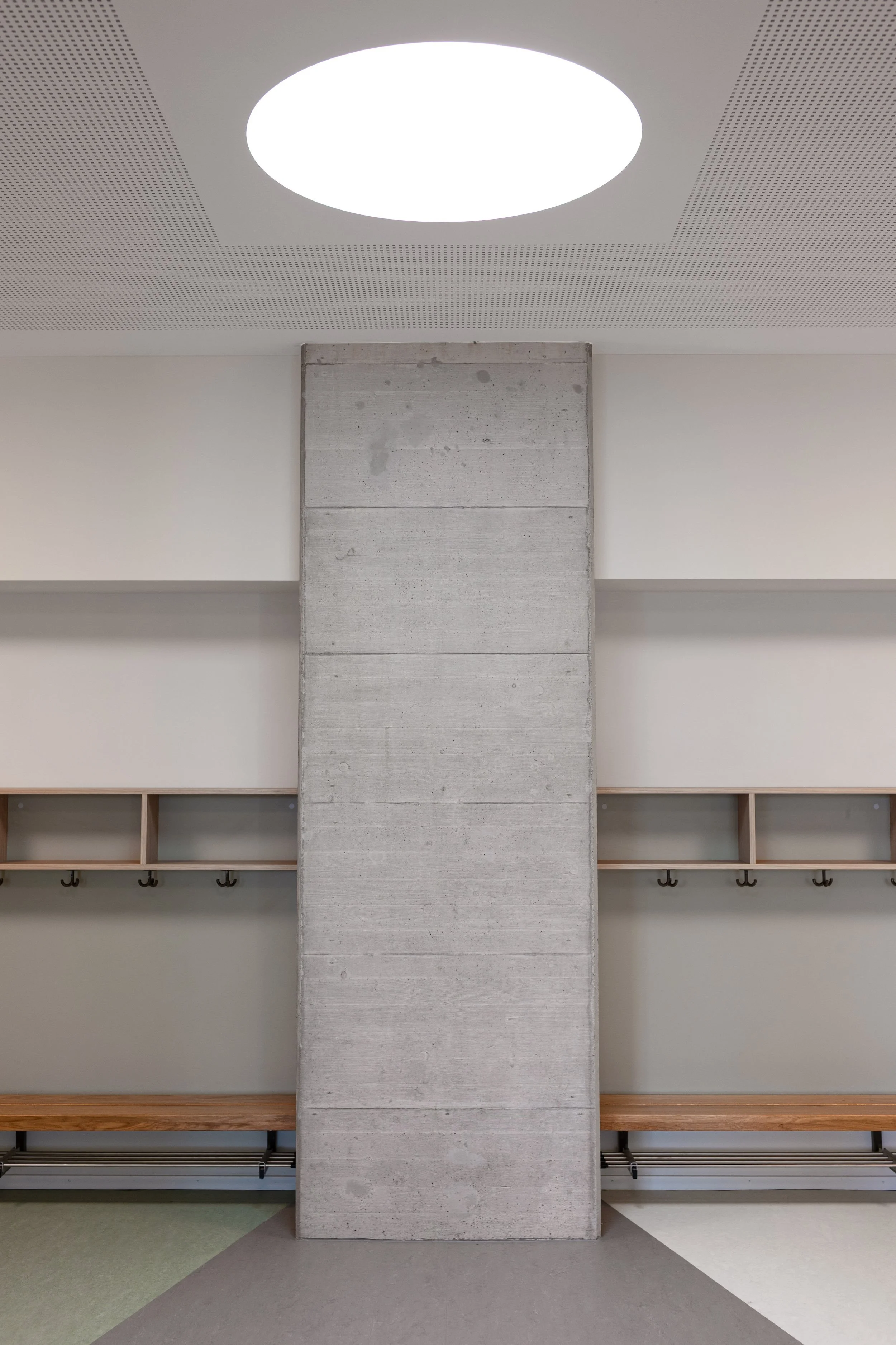 Symmetrisches Detail der Garderobe mit zentralem Sichtbeton-Pfeiler, Holzbänken und rundem Oberlicht. Minimalistische Innenarchitektur.
