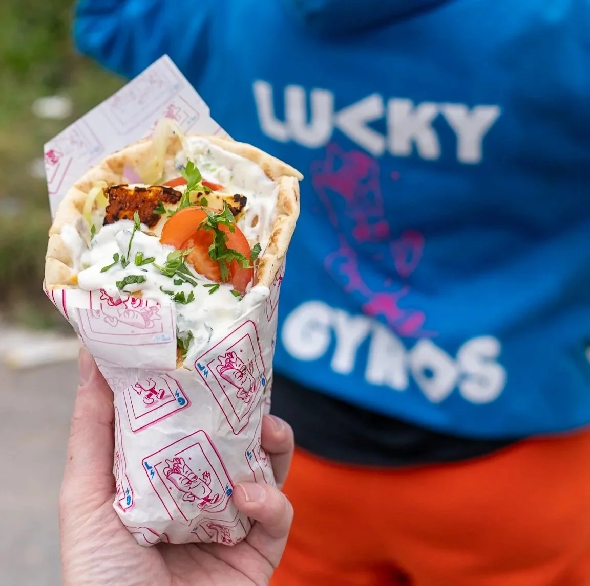Hand holding a gyros with halloumi, tomato, lettuce, red onion, and tzatziki sauce, wrapped in Lucky Gyros branded paper, with a person wearing a blue Lucky Gyros hoodie in the background