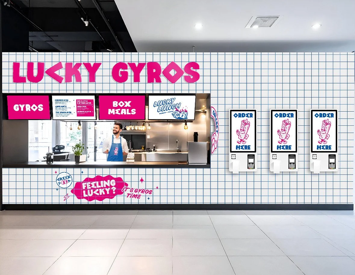 A fast food kiosk called 'Lucky Gyros' with a pink and white color scheme. The signage includes 'Gyros', 'Box Meals', and an animated 'Lucky Lunch' sign. There are three order kiosks on the right side of the wall. A man in a apron stands behind the counter smiling. The wall has a grid pattern with pink text and graphics.