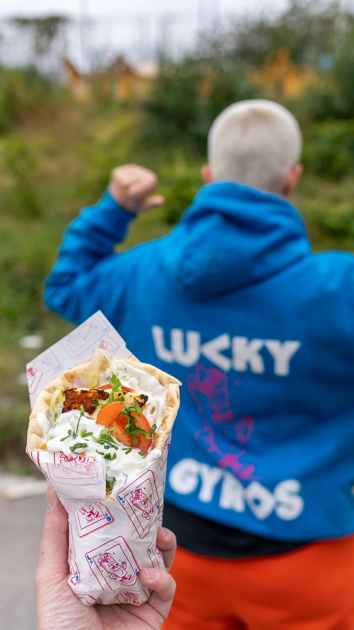 A hand holding a shawarma wrap with salad, tomato, and sauce in front of a person wearing a blue hoodie with 'LUCKY GYROS' on the back, making a fist with a view of the back of their shaved head, outdoors with greenery in the background.