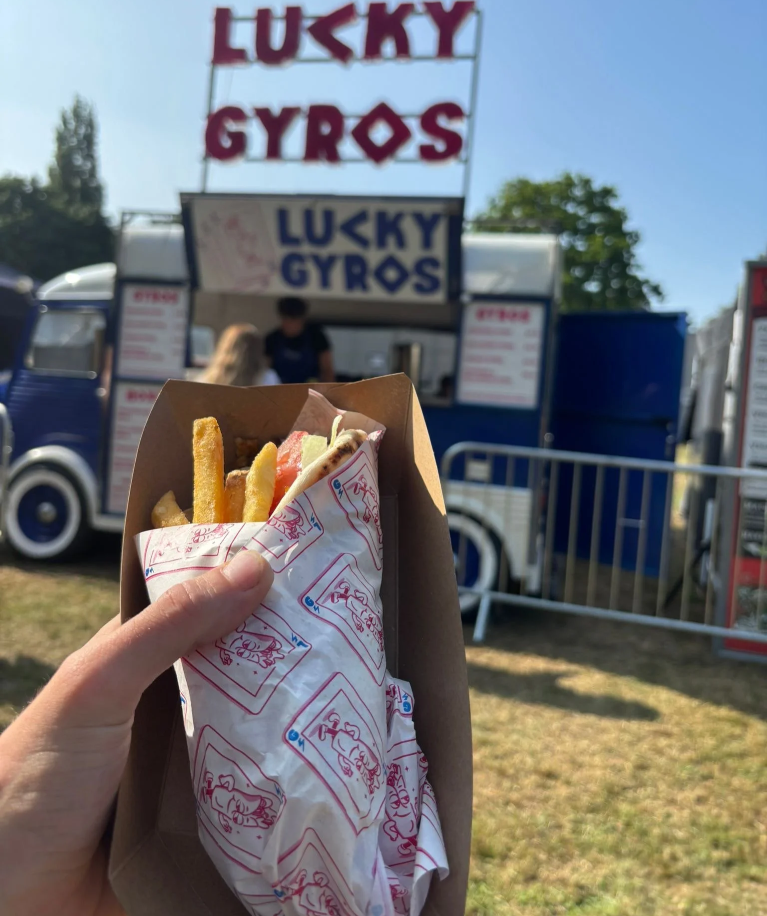 A hand holding a gyros pita from Lucky Gyros, wrapped in Lucky Gyros branded paper. Stood infront of a Lucky Gyros branded food truck at a festival.