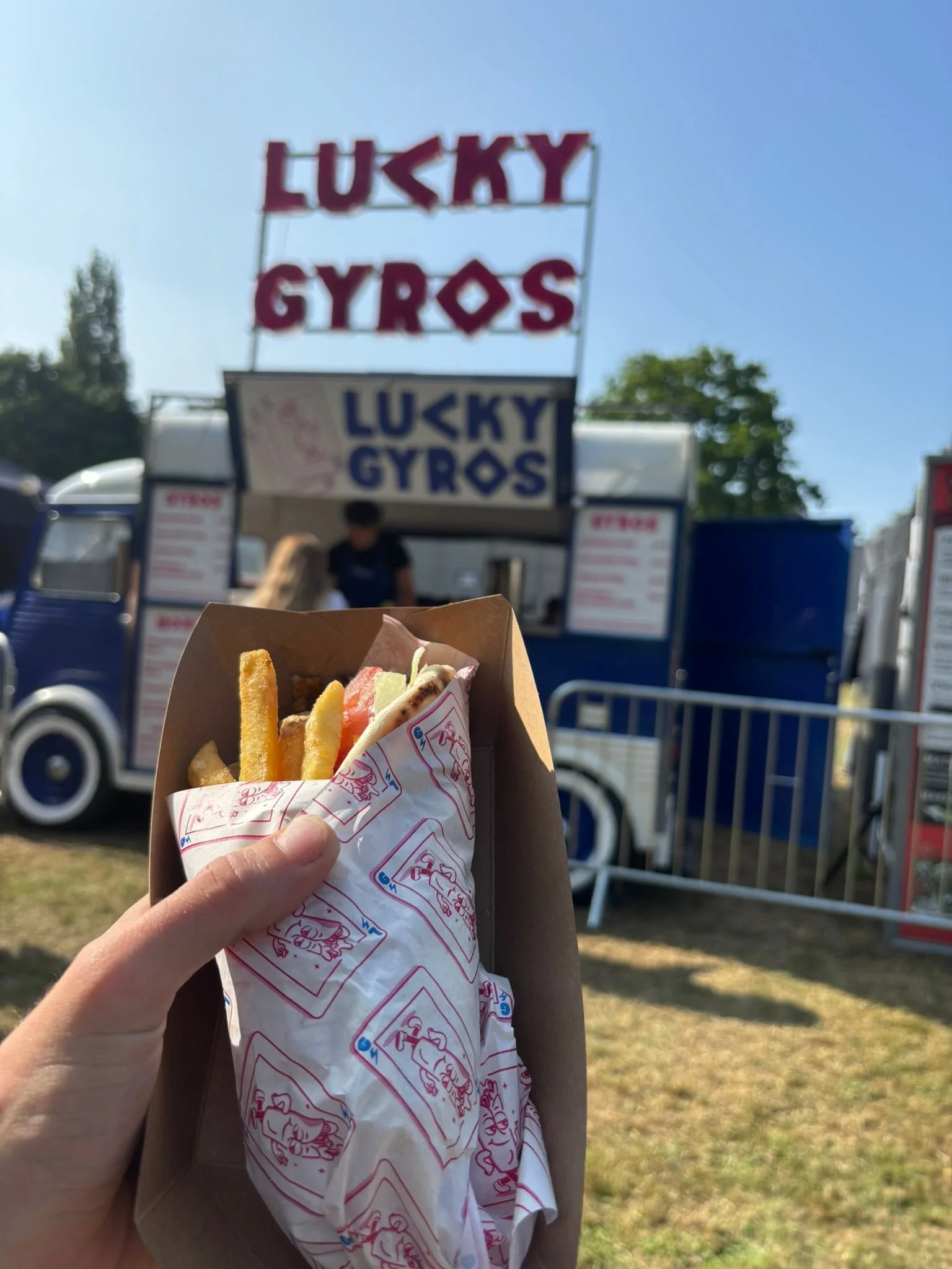 A hand holding a paper-wrapped gyro with fries at an outdoor food stall named "Lucky Gyros" on a sunny day.