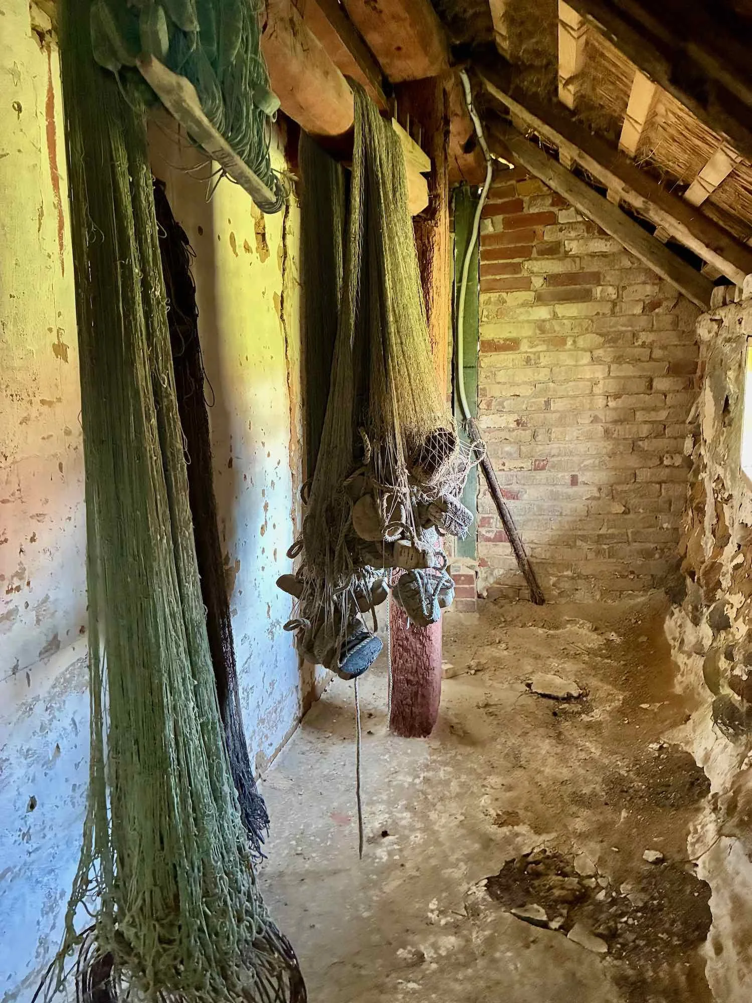 Høstede fiskegarn hænger i en gammel sten- og trækonstruktion, der ligner en lade eller lade. Gulve og vægge ser slidte ud, med sand og snavs på jorden.