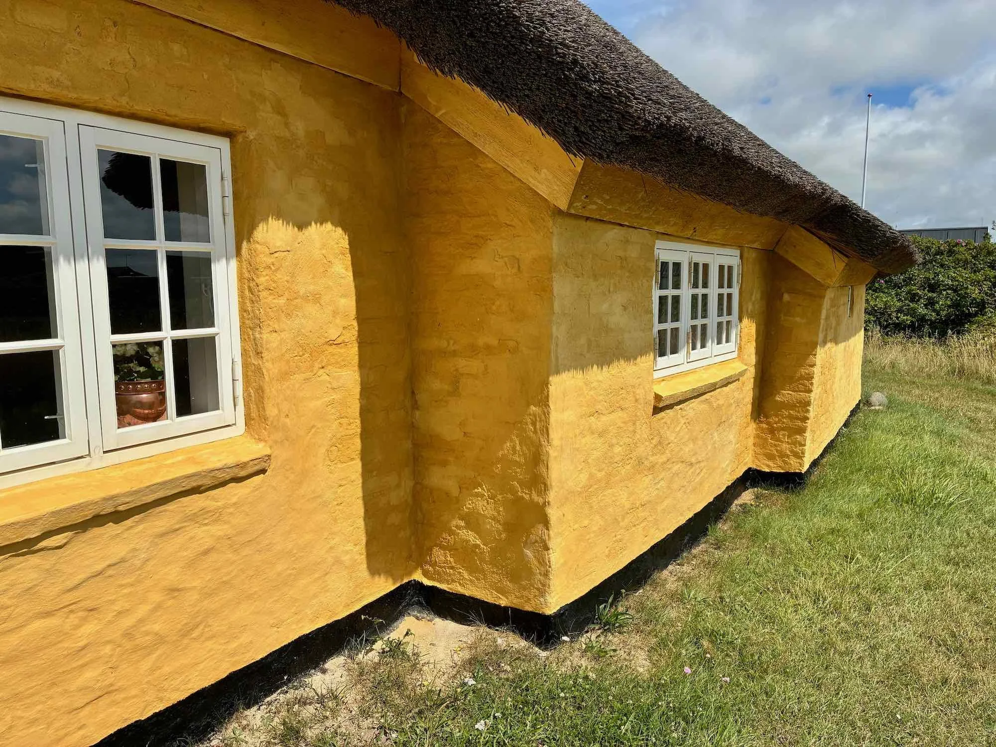 Gult hus med hvidrammede vinduer og stråtag, omgivet af græs.