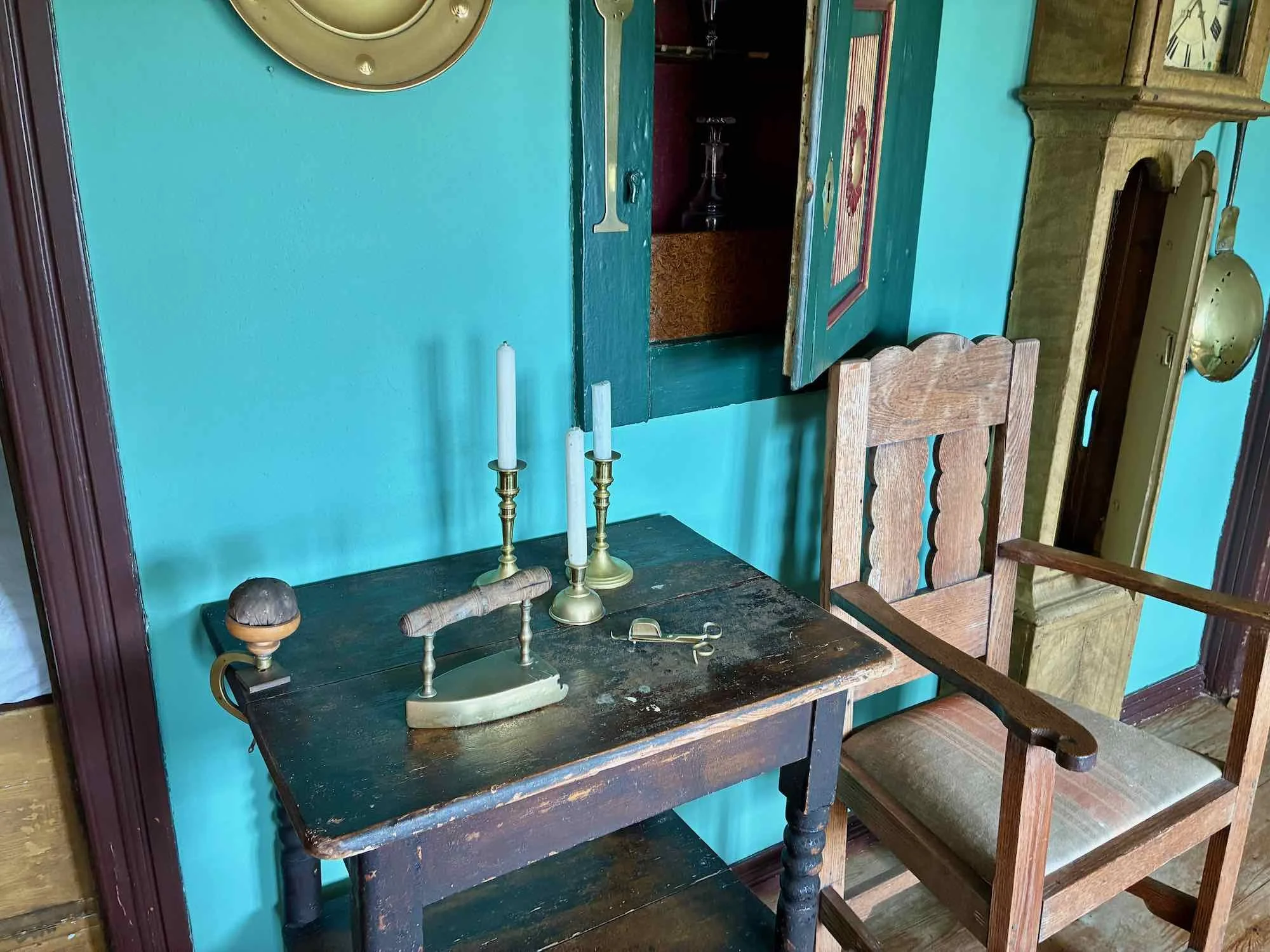 Et gammelt træbord med tre hvide stearinlys i messingholdere, en lille saks og en drejestang, med en træstol ved siden af, foran en blå væg med et vindue og en gammel vægur.