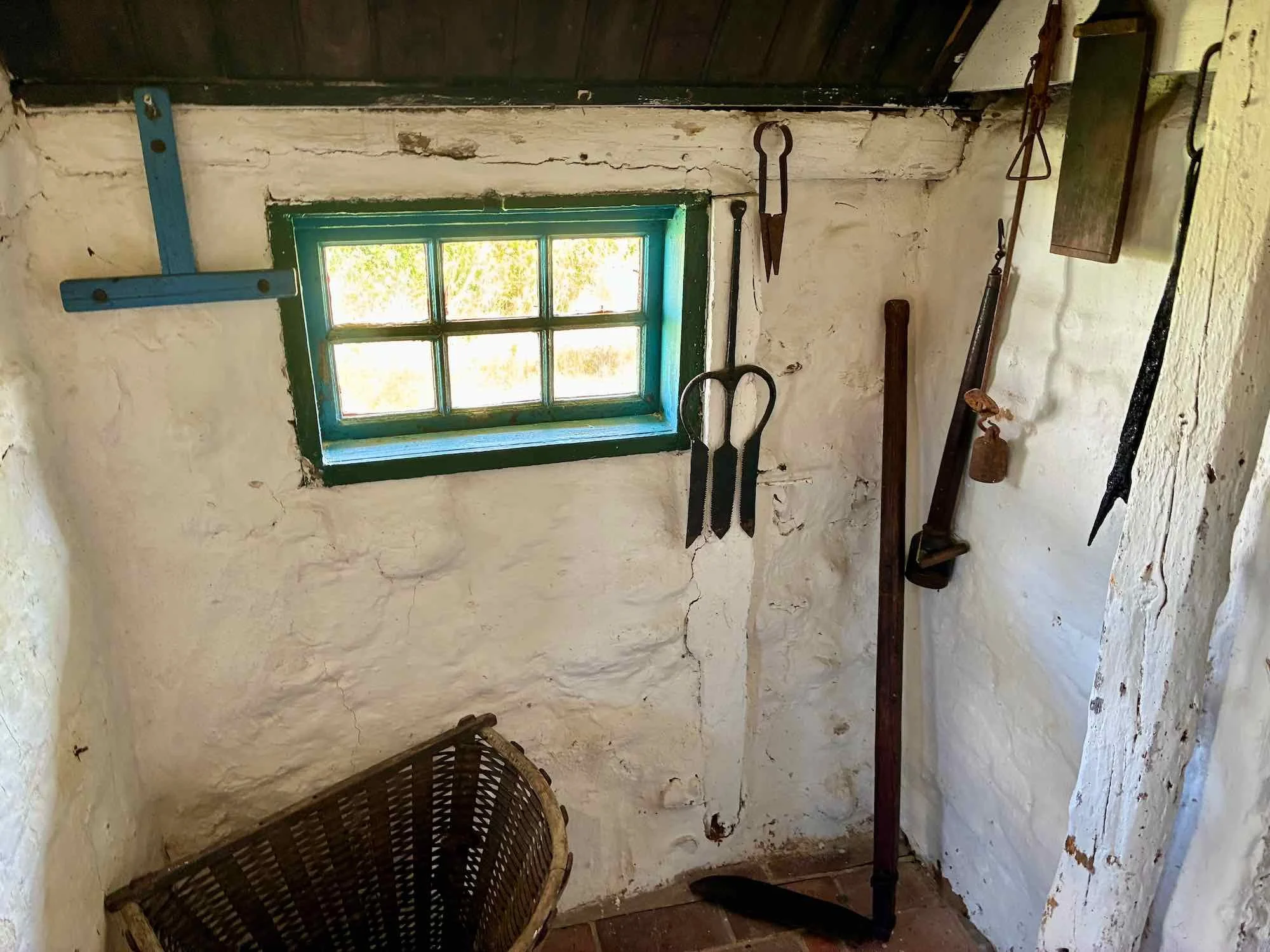 En ældre lukket stue med små vinduer, væggene er hvide og rå, og der er gamle landbrugsværktøjer hængende på væggen.