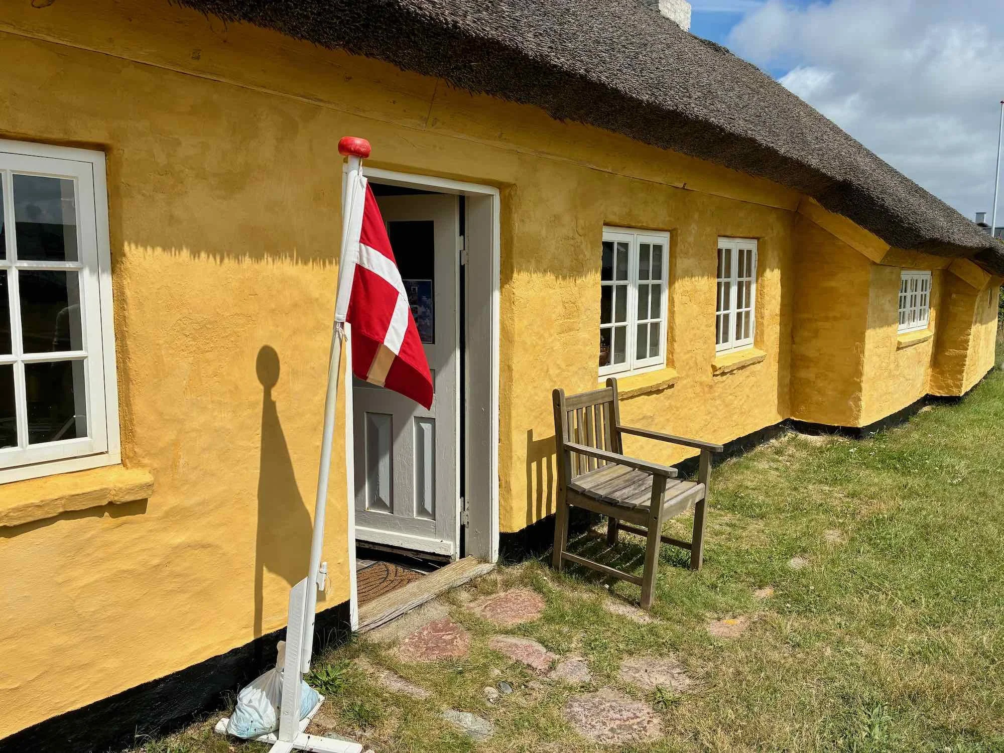 Det gamle fiskerhus i Agger med hvide vinduer og en dør, der er åben. Der står en skille med et dansk flag foran døren og en træbænk ved siden af døren på græsplænen.
