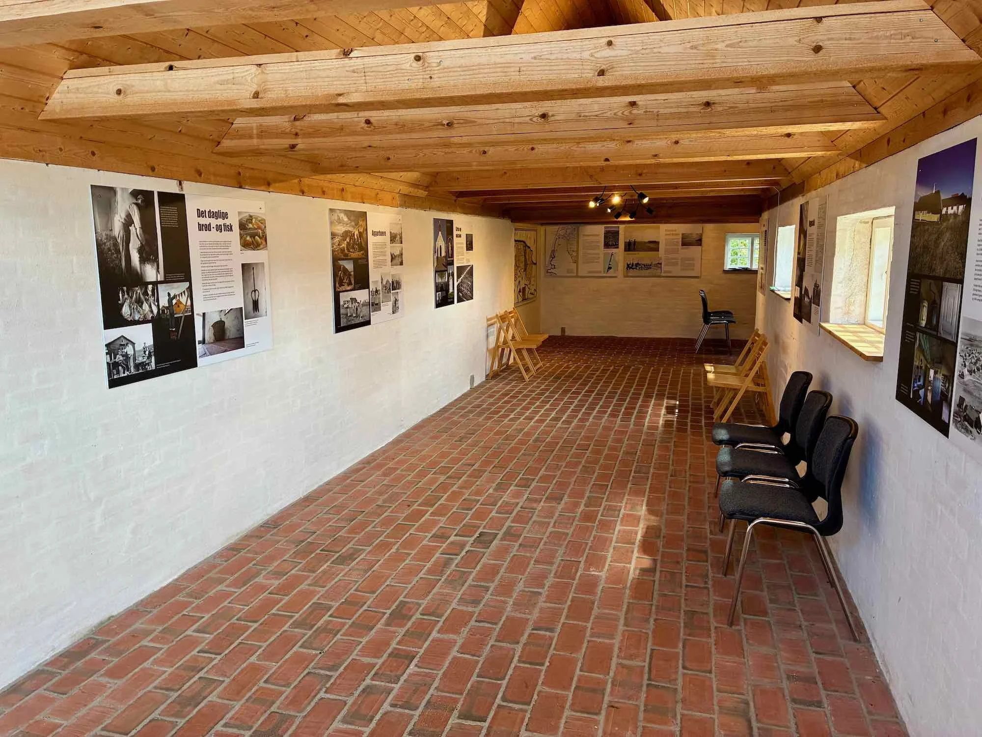 Indendørs udstilling med fotografier og tekster på væggene, flere sorte stole langs væggen, træloft og gulv af røde mursten, små vinduer og nogle træstole i hjørnet.