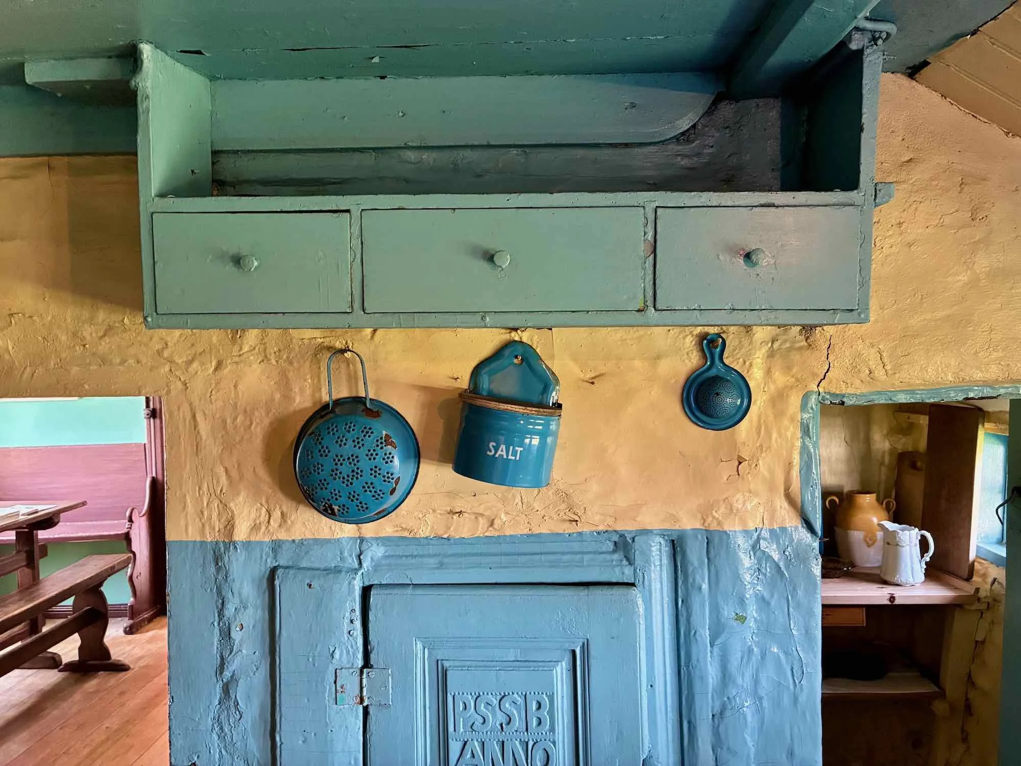 Interiør af et gammelt køkken med blå og gul væg, små skuffer i en grøn skab, og blå køkkengrej hængende, herunder en saltkande og en skål.