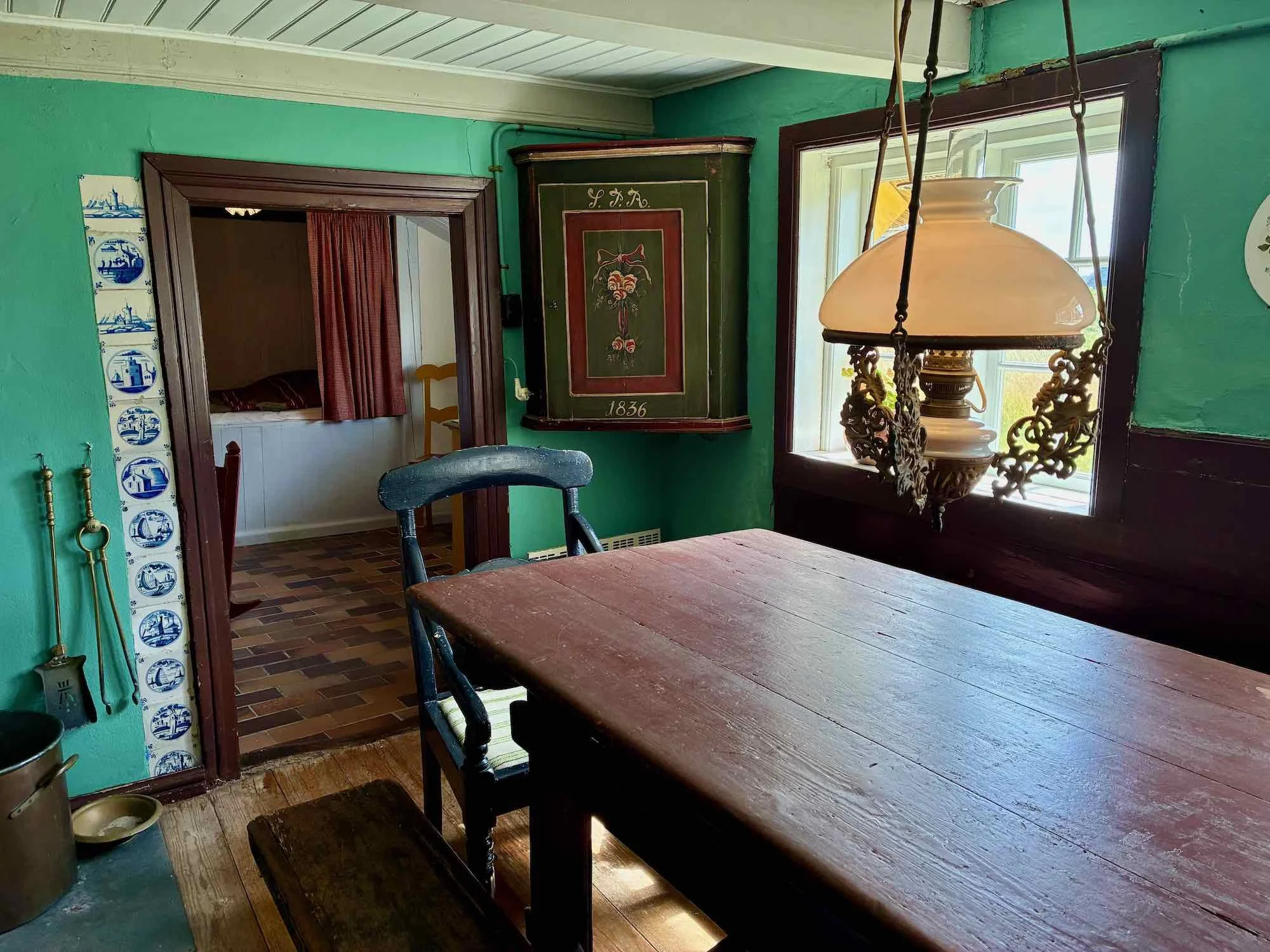 Et gammelt køkken med grøn væg, et træbord, historisk lampe, ramme med blomster, vindue med udsigt, og en dør, der fører til et andet rum.